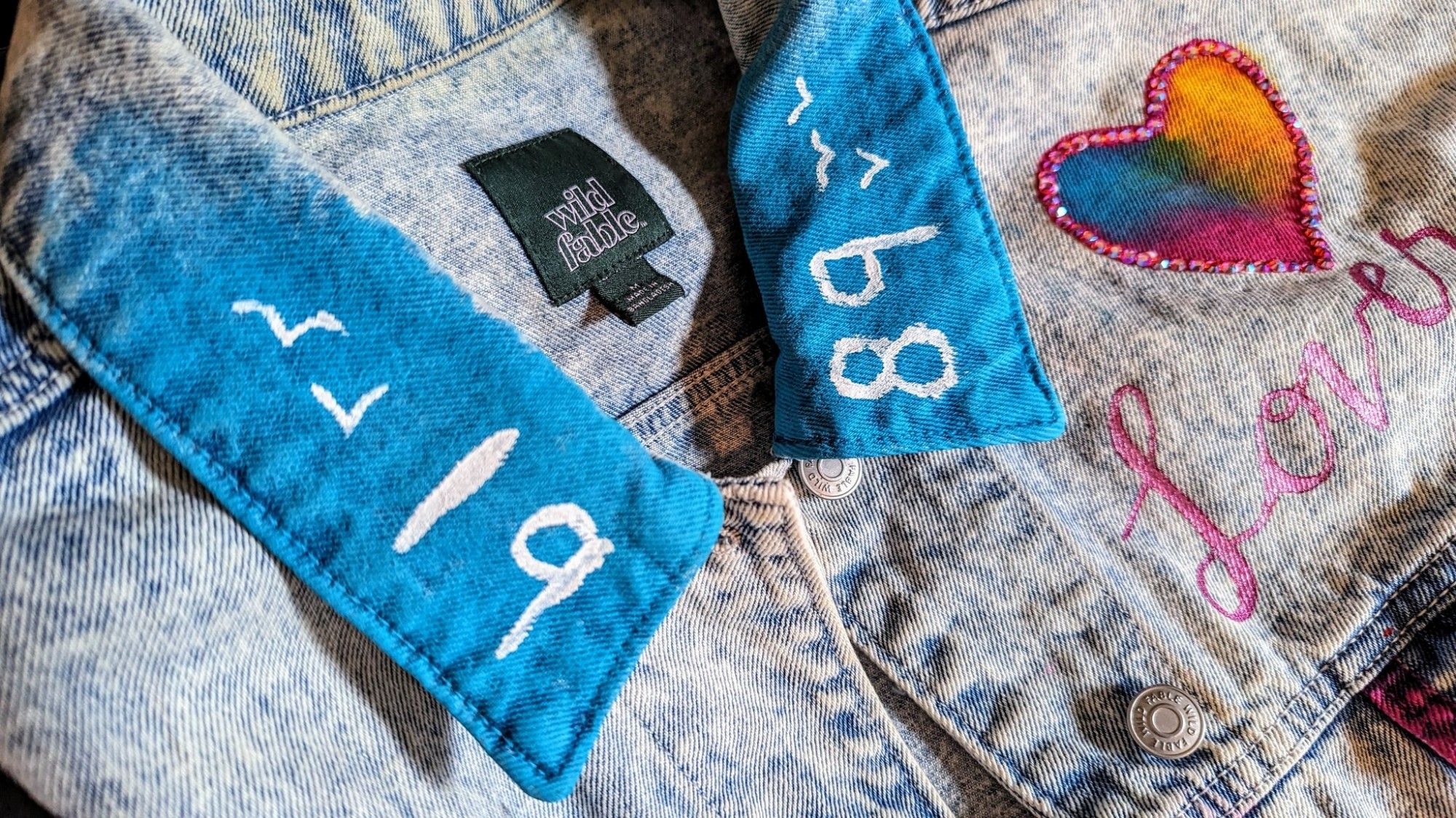 Close up photograph of a light wash denim jacket. The collar is painted a bright blue that fades out towards the back. On one side of the collar there are two small, stylized birds and the numbers "19" painted in white. On the other side there are the numbers "89" and three stylized birds, also in white. In the background, over the shoulder and upper chest section of the jacket, is the word "Lover" in script, painted in pearlecent pink paint. There's a pink, blue, and yellow heart above the word that has a border of pink rhinestones.