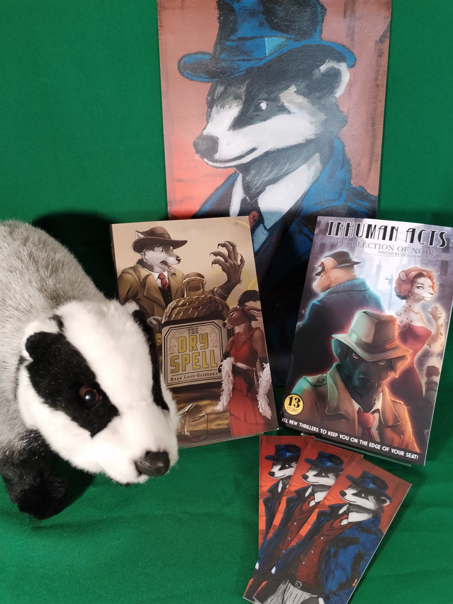 A copy of the book 'The Dry Spell' by Ryan Loup-Glissant, a copy of the book 'Inhuman Acts' edited by Ocean Tigrox, a plush badger, and a banner and bookmarks showing a badger dressed as a noir-style detective