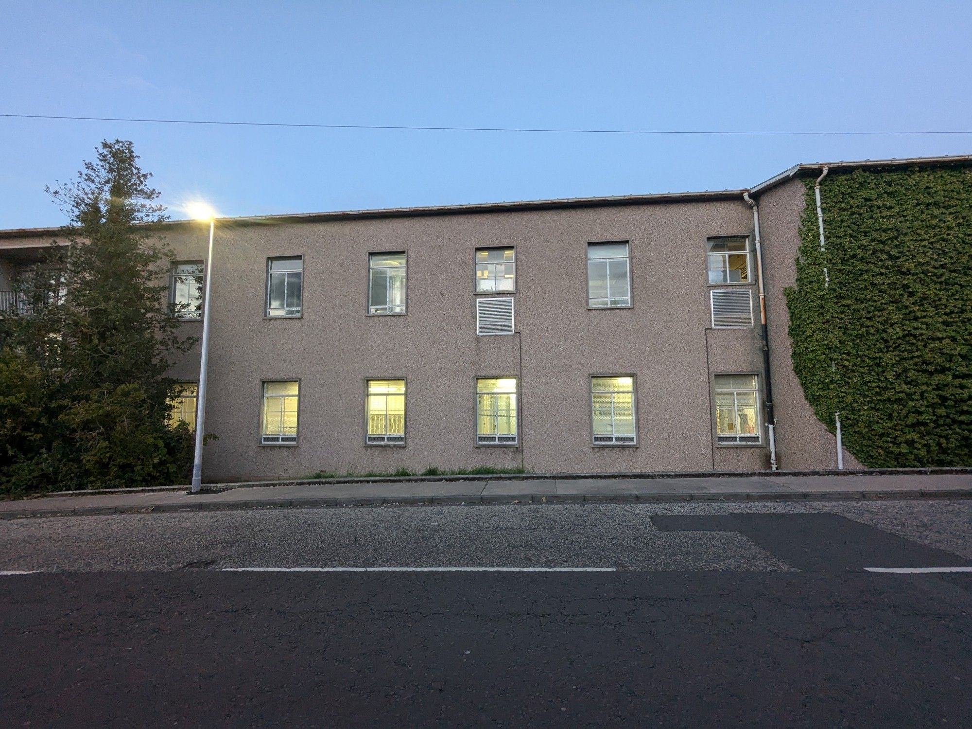 A two storey 1950s telephone exchange building. Its lower windows are lit from within. A creeper is growing up its rough cast walls. The sky is blue and dusky. A streetlight has just come on.