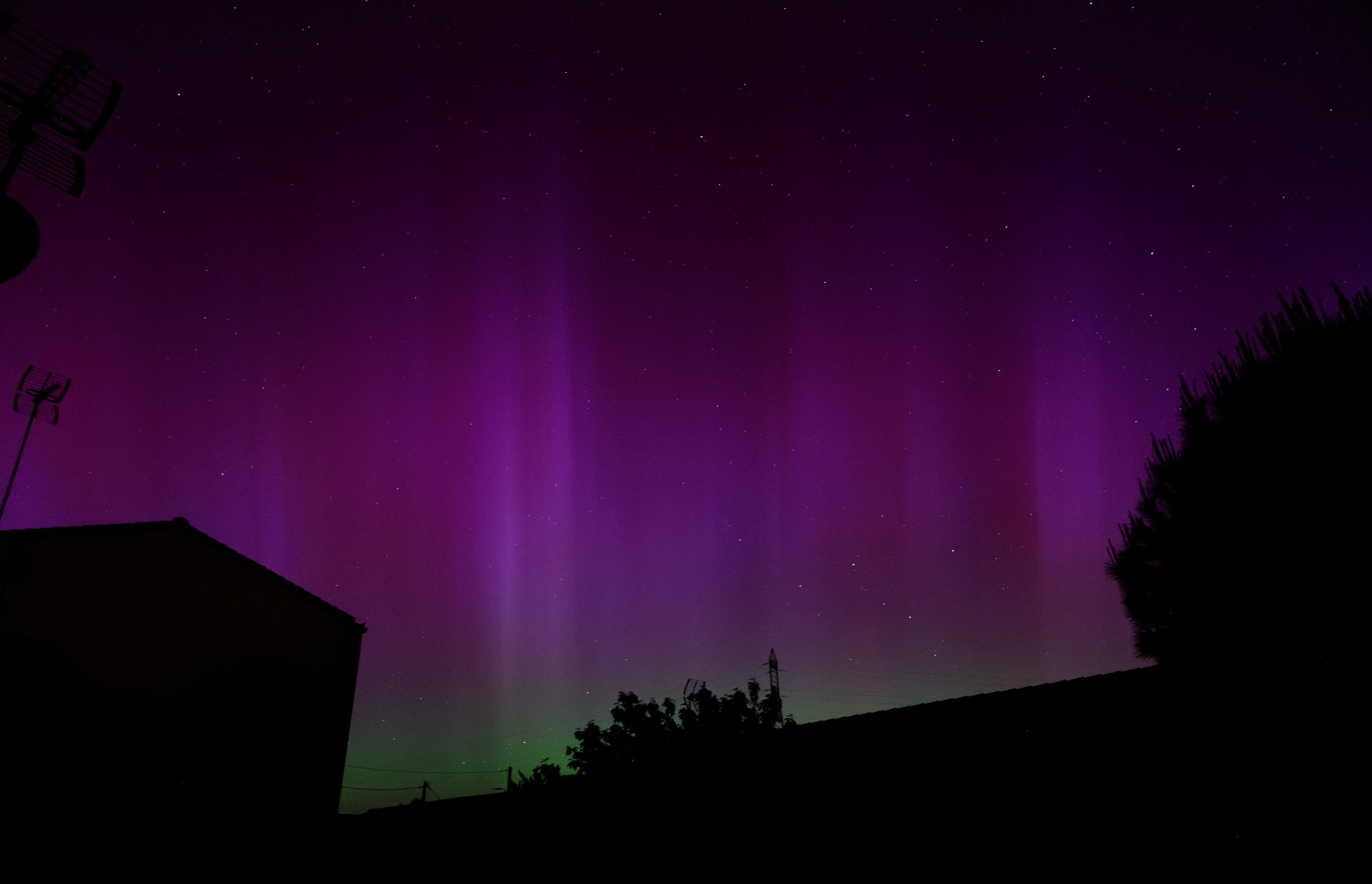 Photo d'une aurore boréale prise depuis mon jardin en mai 2024
Dégradé de bas en haut de vert et violet sur silhouette de maisons et d'arbres 
Le ciel est strié de rayons verticaux dans les mêmes tons que l'aurore, mais plus clairs
(Ça scintillait, ce que ne rend pas la photo)