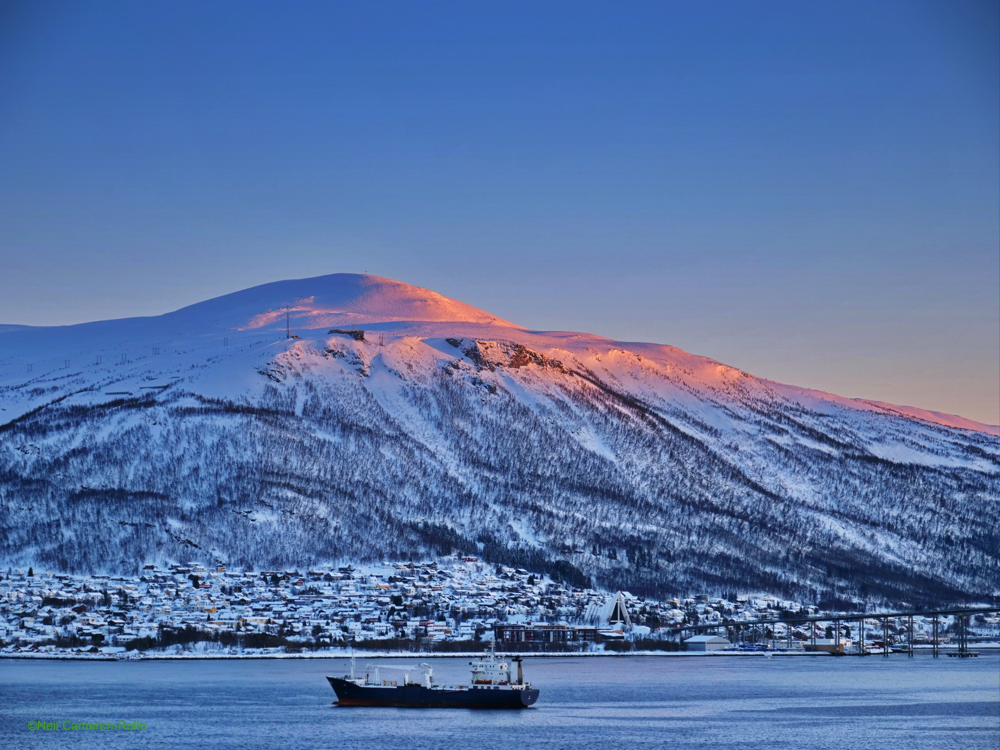 Shot towards the South East from Tromsø island. Across the water Tromsdalen (a suburb of Tromsø) hunkers beneath a tree clad, snow covered mountain. The setting sun catches the top and edge of the mountain turning it gold and pink.
The triangular structure in Tromsdalen in the Arctic Cathedral, high up on the mountain the building of the Tromsø Obsevation point can be made out (it is accessible by cable car), if you can zoom in enough you will see people standing on the ridge beside it.