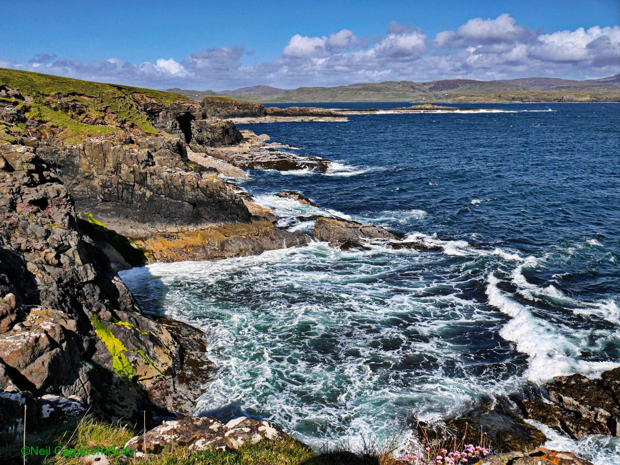 Taken on Oronsay Island in the direction of Ardreck Point (roughly North-North East).
The rock shore of the island leads away from the camera (on the left of the image), low cliffs are battered by the blue, wind driven waters of the North Atlantic, there is a wave cut cave on the middle distance.
Across the wave tossed water the low hills of the West side of Skye stand up against the blue sky and have a  a cap of white fluffy clouds.