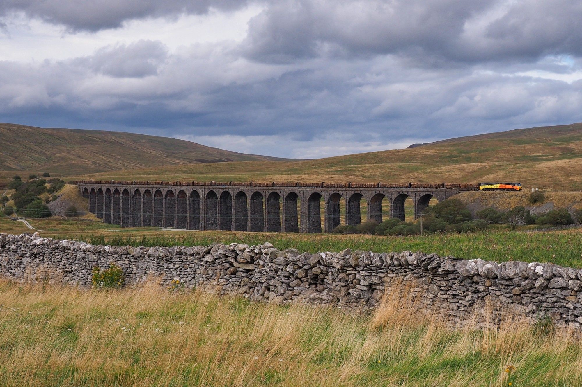 The same freight train as before passing over a massive, long viaduct in an empty moor landscape. Stone walls and dry grass in the foreground. Cloudy sky.