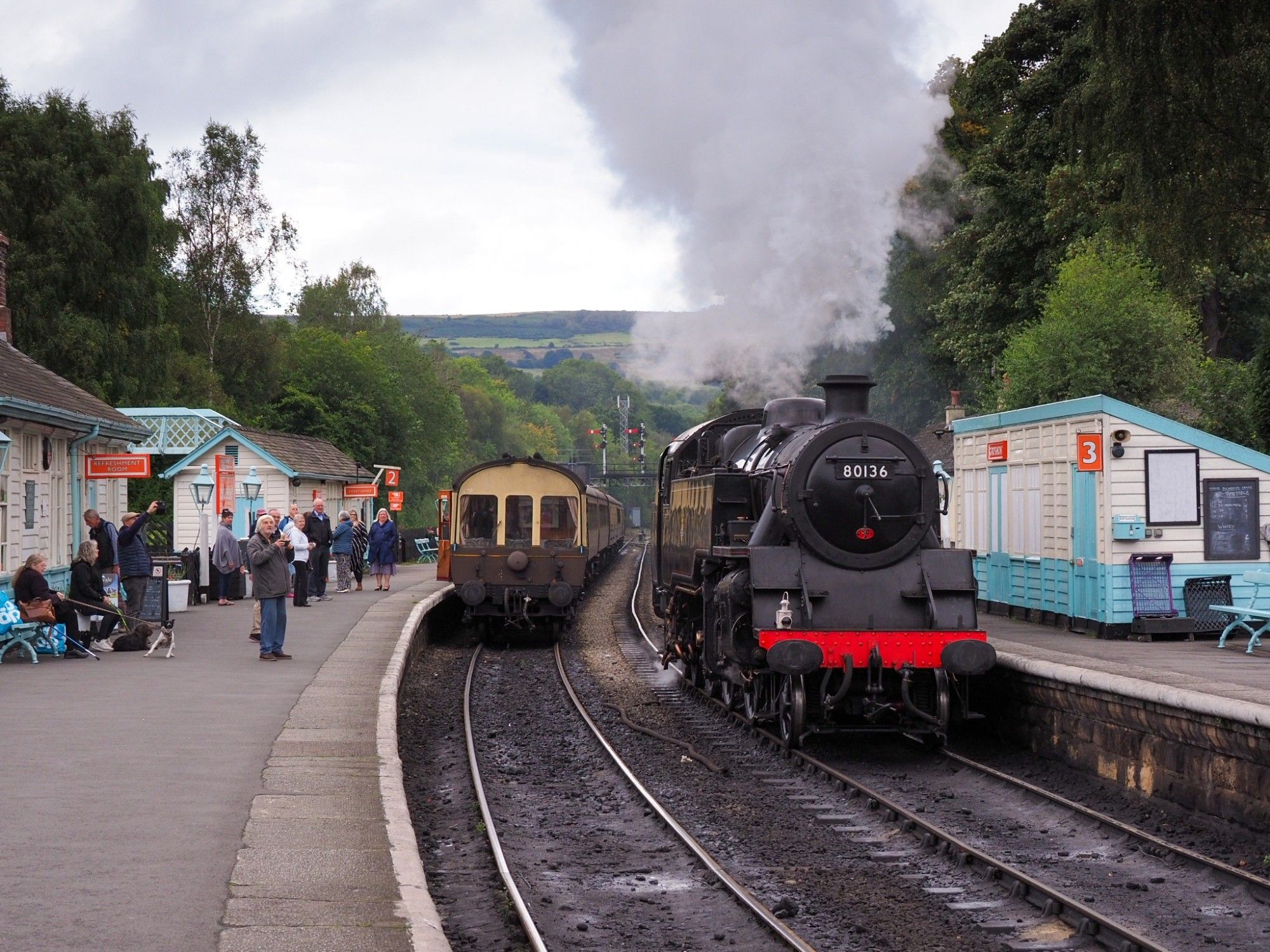 Steam loco running around a rake of carriages. Wooden buildings on the platforms