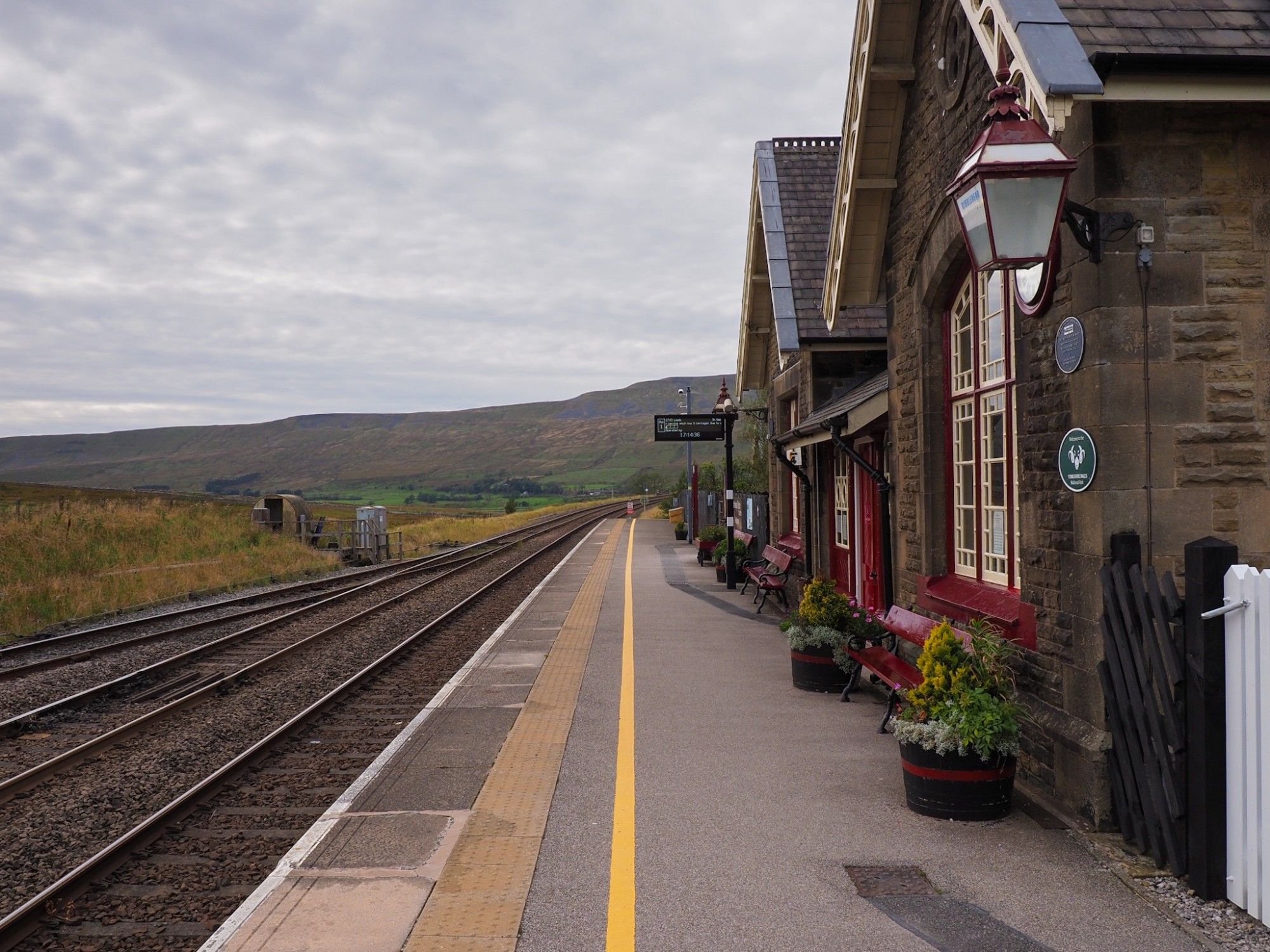 Beautiful stone building of Ribblehead station, platform side. Flowers in pots and wooden benches on the platform.