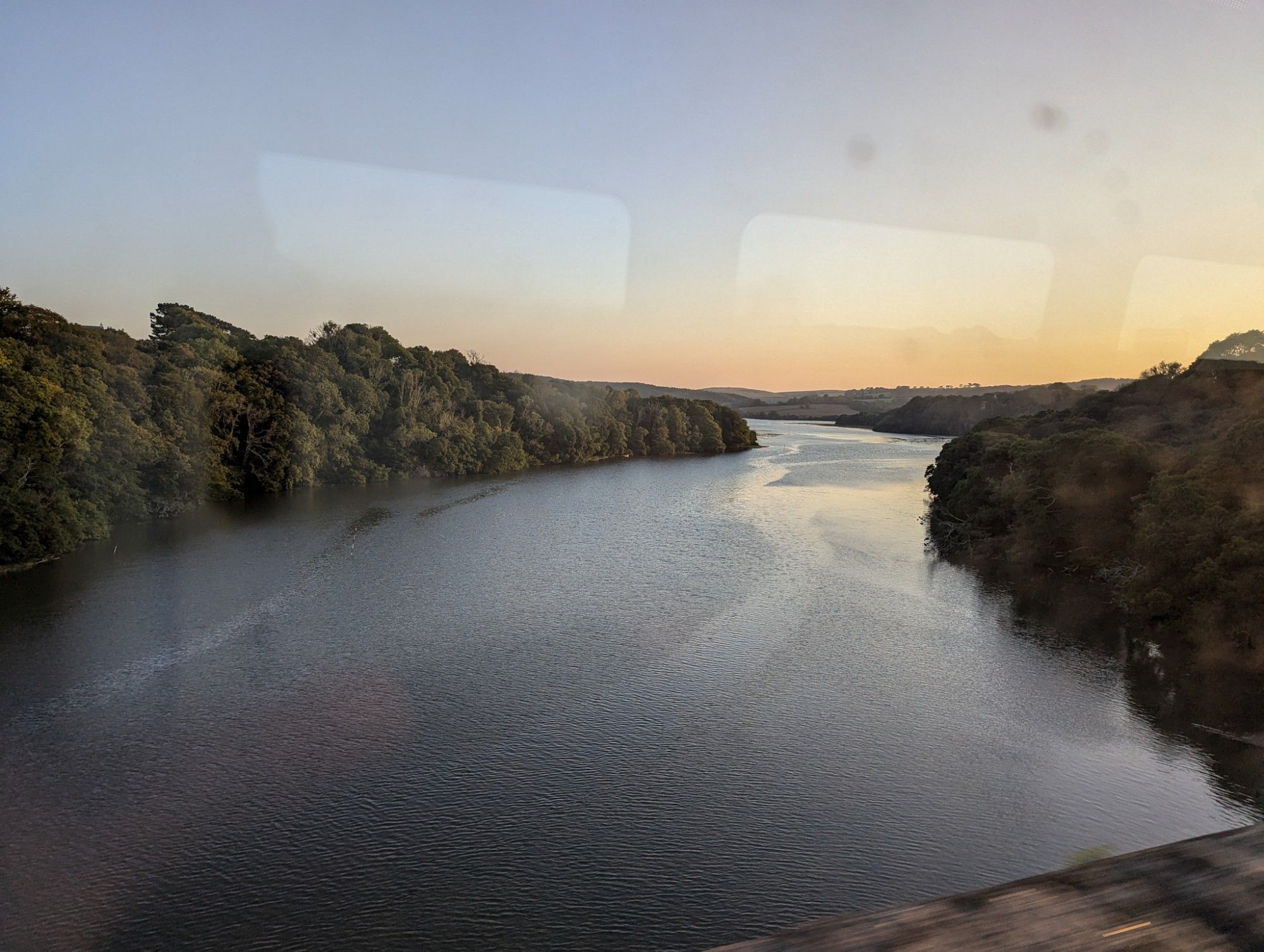 View through the train window: The line crosses a body of water which looks like a lake, but is actually a river near its estuary. Trees to both sides, sunset colours in the sky.