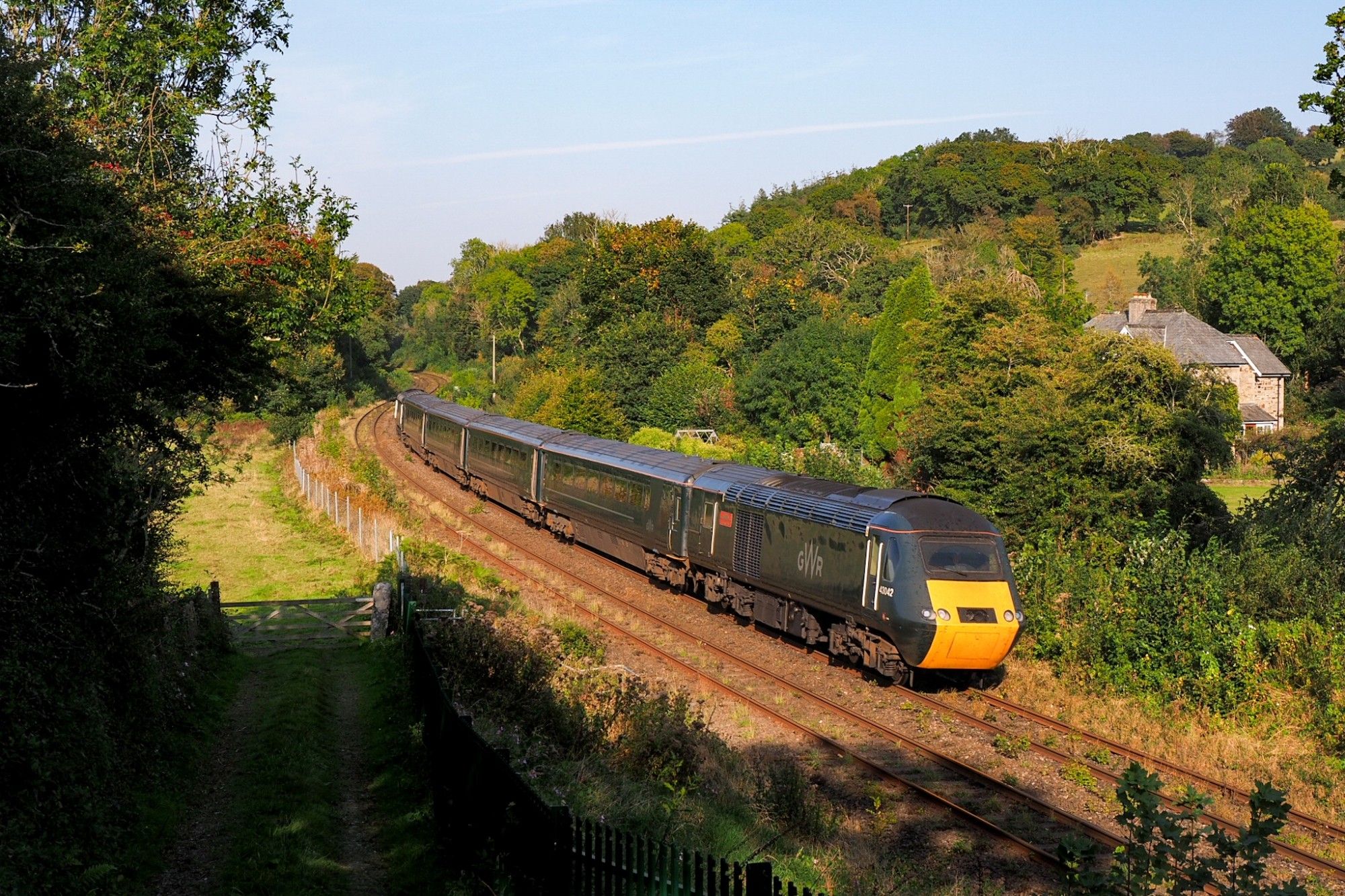 Another GWR HST rushing through a curve in a green valley, seen from a slightly elevated position on the outer side. Behind, hidden by bushes, a british countryside house. Evening light, tree shadows in the foreground.