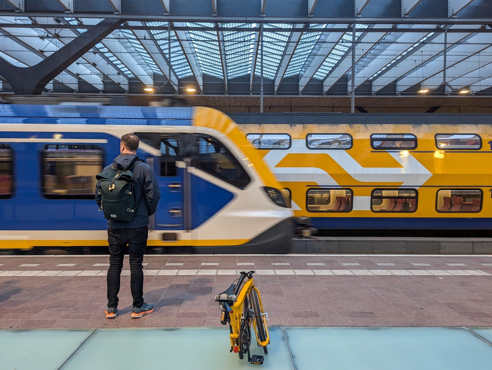 Sideways view on a yellow VIRM train standing in Rotterdam-Centraal. A blue sprinter is just pulling in (slightly blurry). A passenger and my bike are waiting in the foreground.