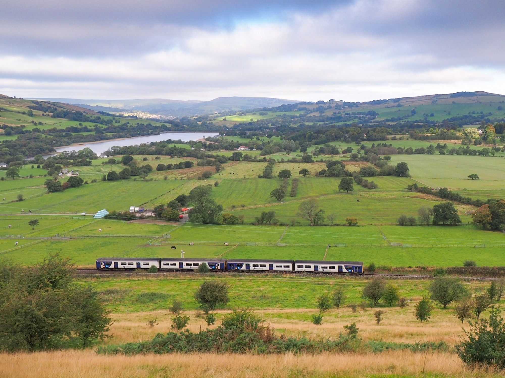 Panorama view over a hilly, green landscape with pasture, hedges and a reservoir in the background. Cloudy grey sky with a single blue spot. In the foreground, below the photographer, a double unit class 150 passing.