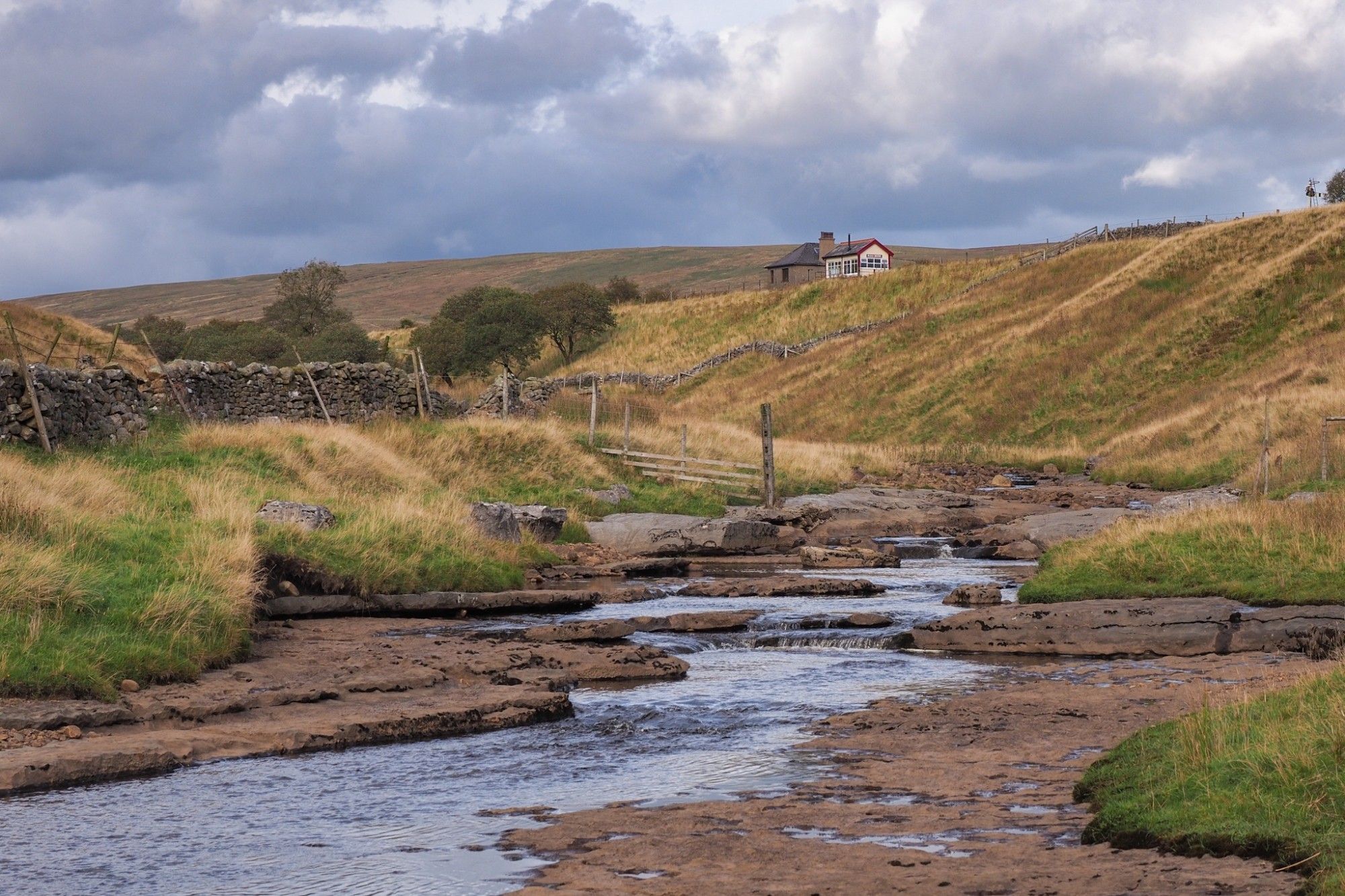 A small river flows over flat rock plateaus in a valley. Behind, up a hill, two buildings.