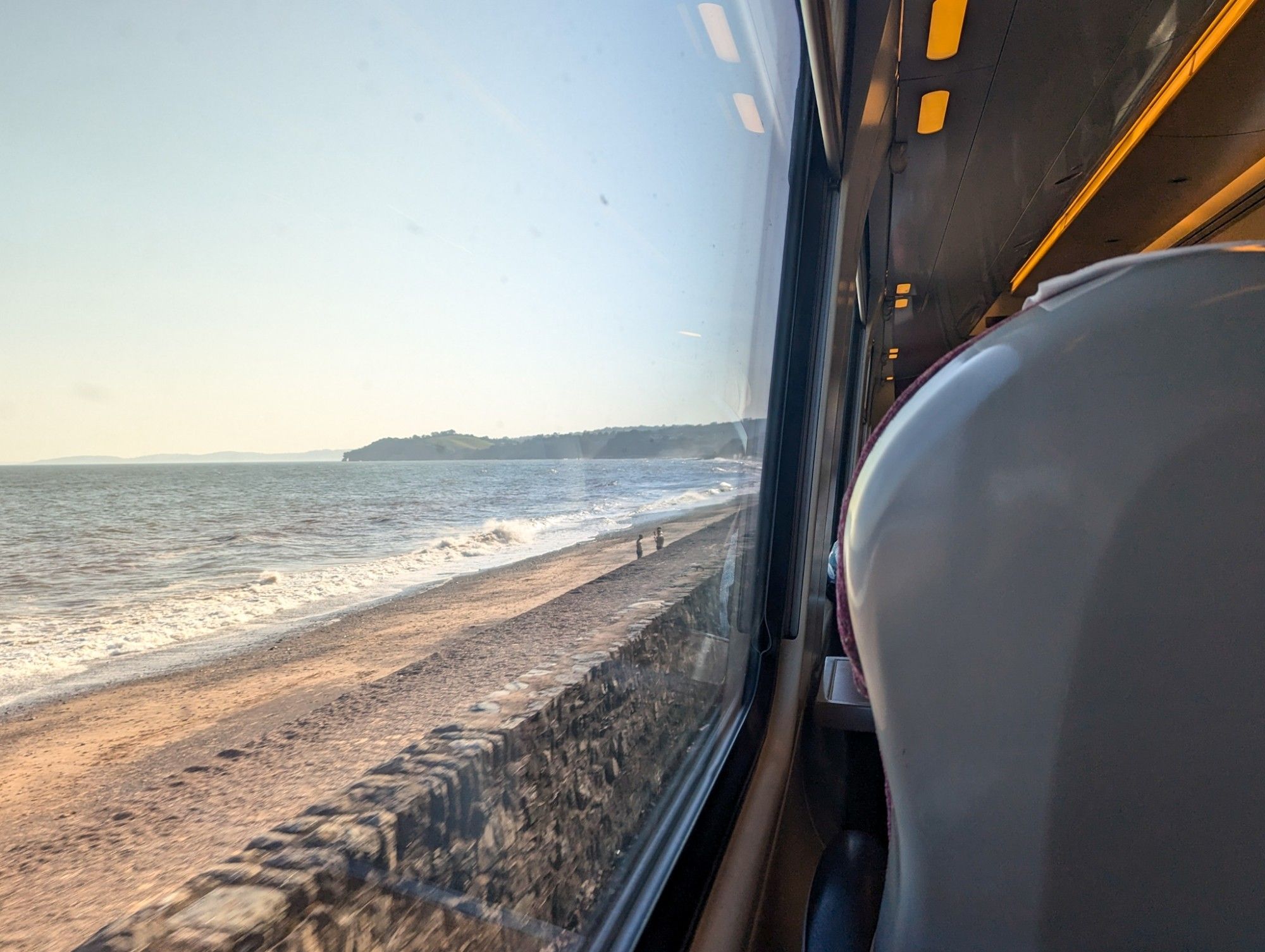 View though the train window: The train drives along the sea, a stone wall, a sandy beach and the sea just next to the tracks.