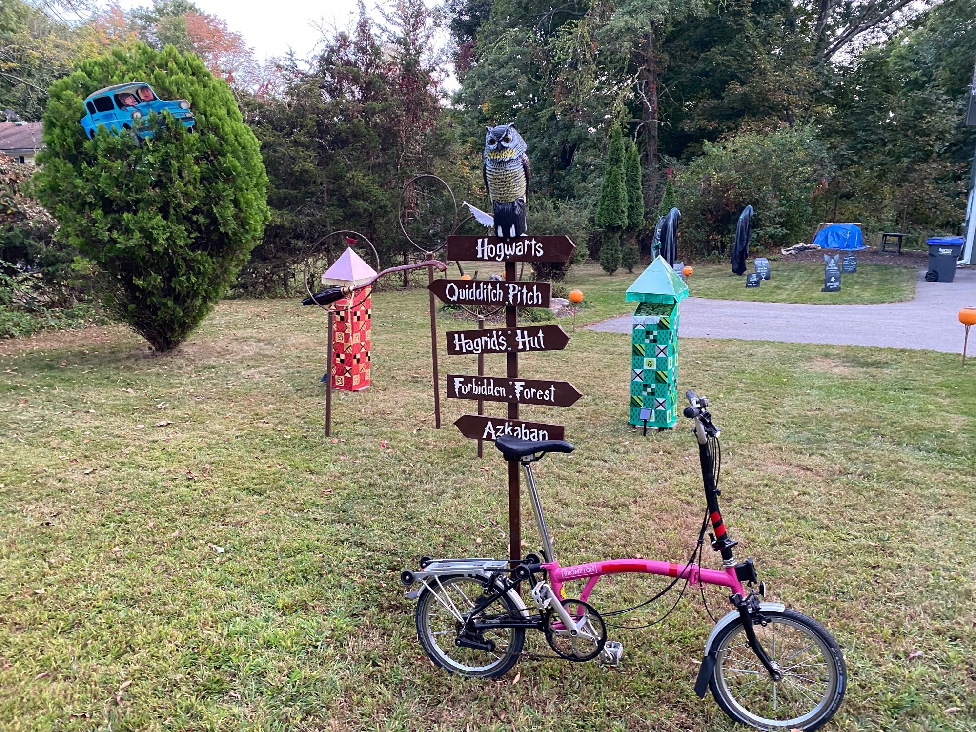 A pink Brompton bicycle leaning against a Halloween decoration Harry Potter themed sign with quidditch towers and a flying Mini car in a tree