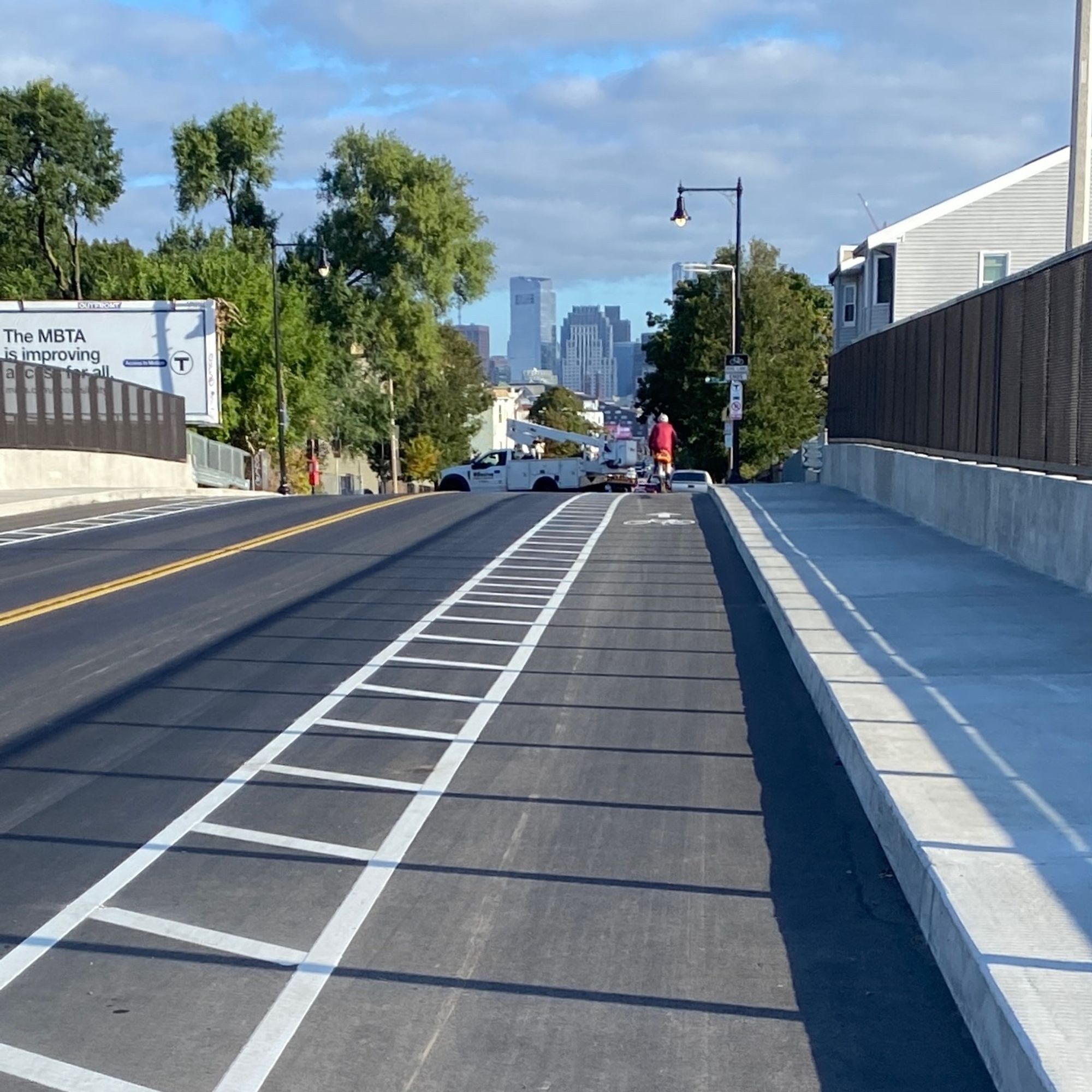 The view from the bike lane of the new Dorchester Avenue bridge, with a sidewalk and fence at the right and downtown Boston in the distance framed by trees and houses