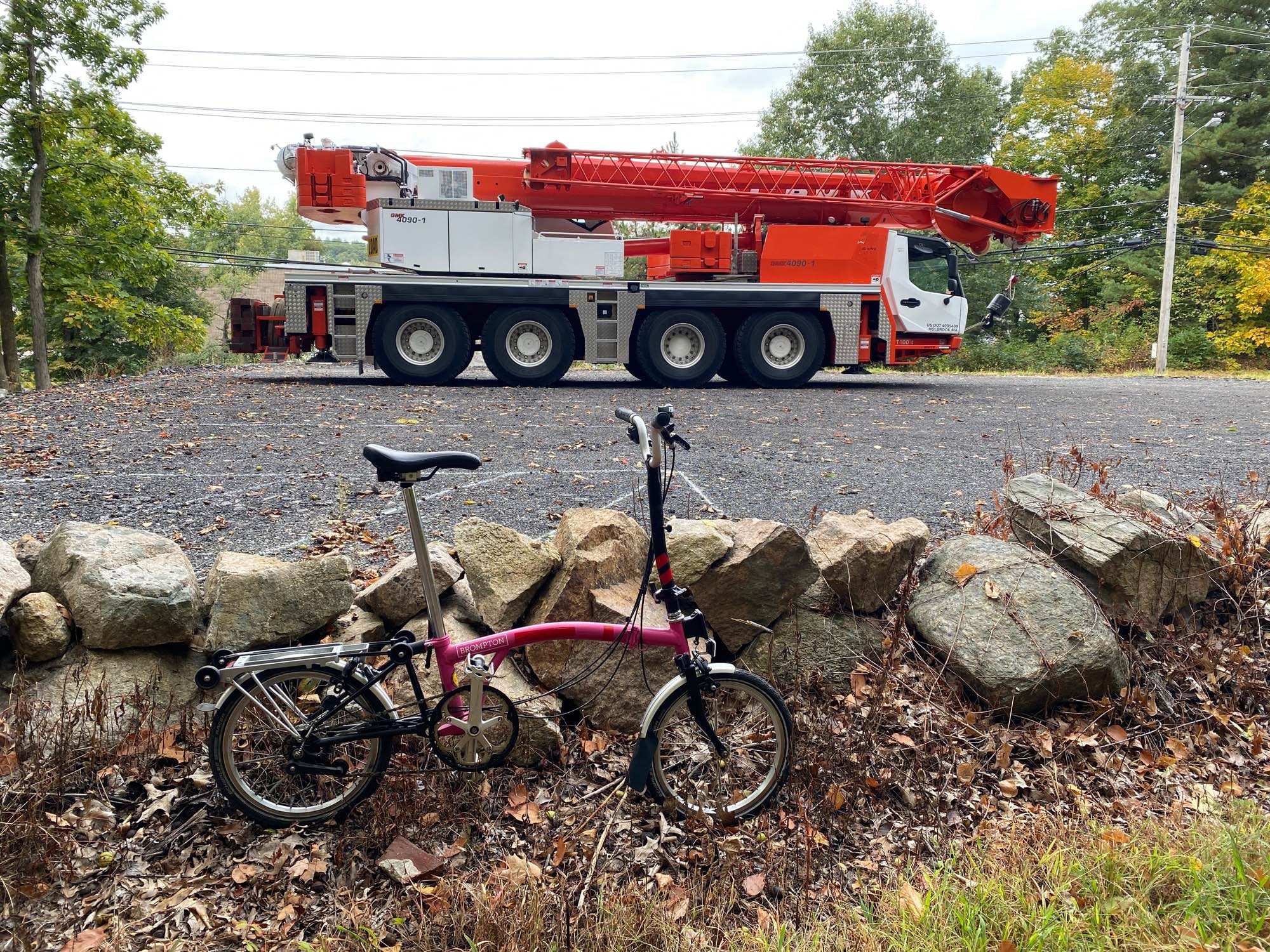 In the foreground a small pink folding Brompton bicycle leaning against a stone wall. In the background is a large orange and white mobile crane truck with four big tires on each side.