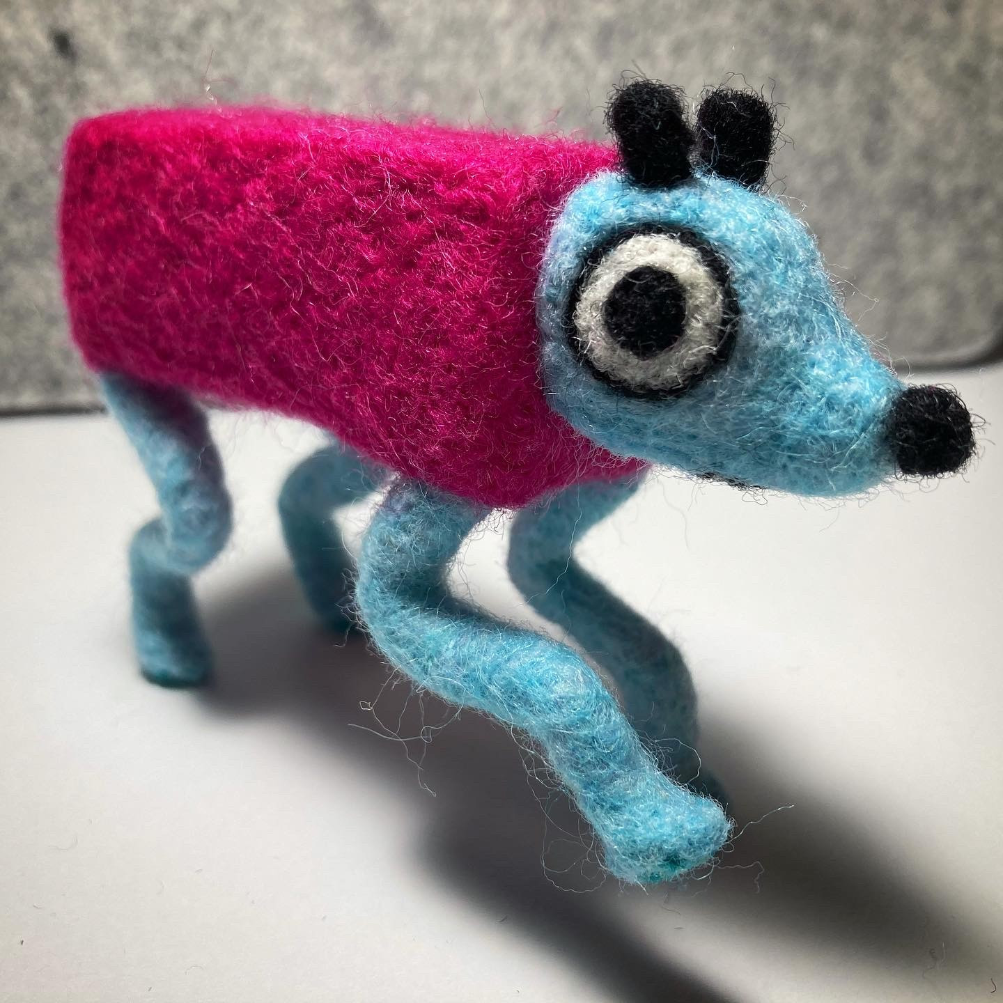 A photo of a needle felted Wobbledog from the game Wobbledogs. It's standing on three legs and taking a step towards the viewer.