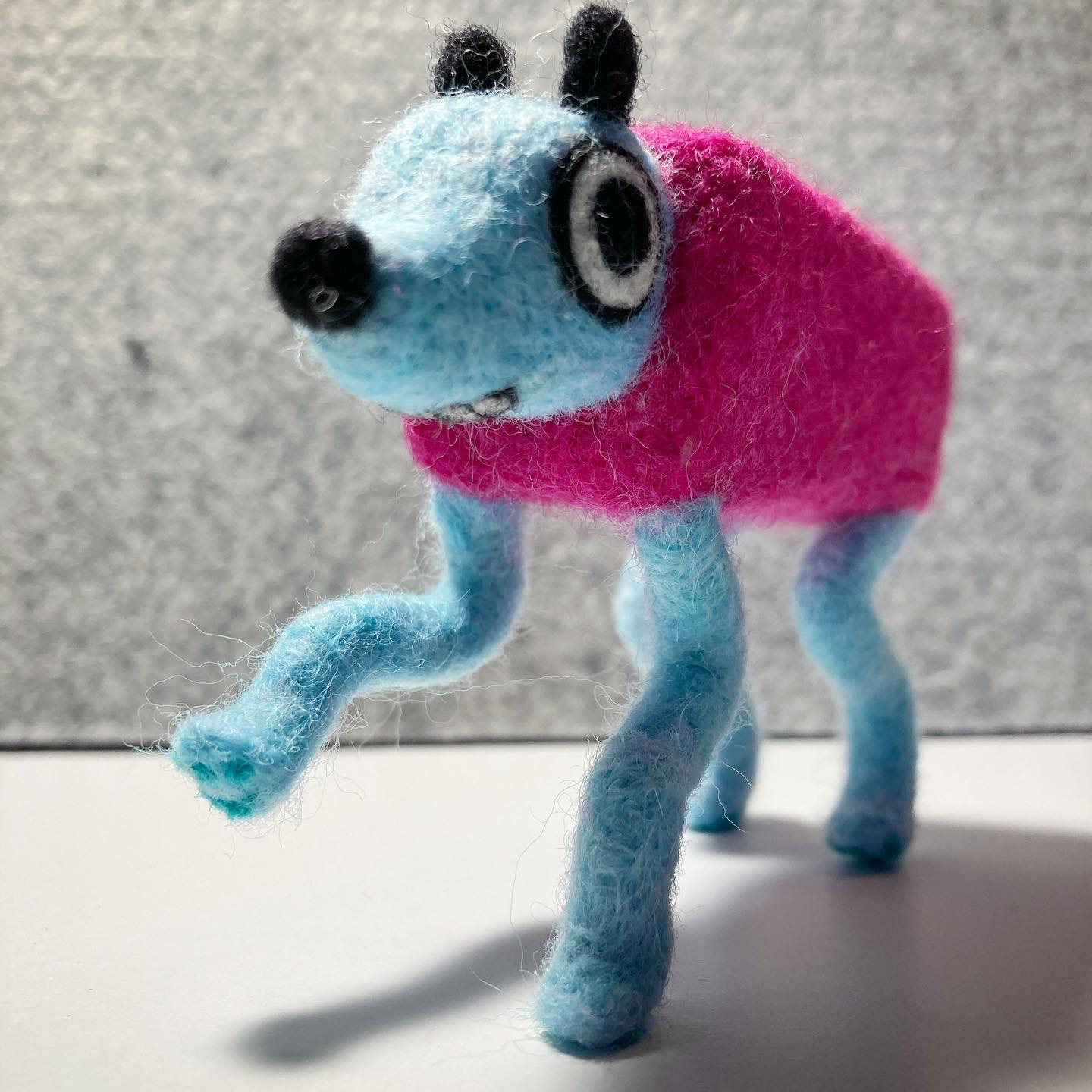 A photo of a needle felted Wobbledog from the game Wobbledogs. It takes a step towards the viewer. It's mouth is slightly visible underneath it's head.