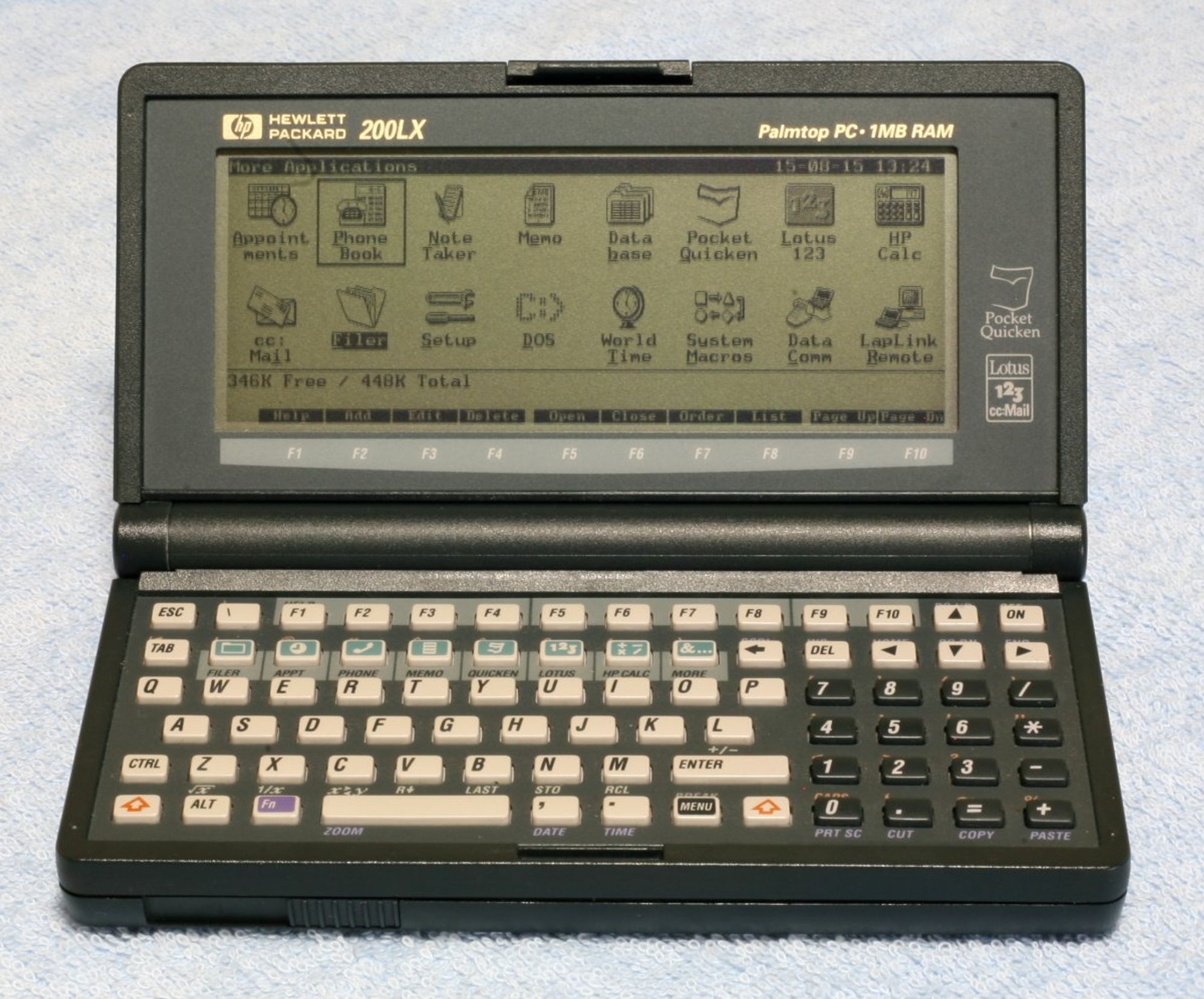 A grey clamshell handtop.  Up top, the 640x200 monochrome screen has applications:  Appointments, Phonebook, Note Taker, Memo, Database, Pocket Quicken, Lotus 123, HP Calc, cc:Mail, Filer, Setup, DOS, World TIme, System Macros, Data Comm, and Laplink remote, plus a memory tracker (346K Free/448K Total) and legend for the function keys.  Down below, a micro-QWERTY keyboard with numb pad appears, with a second row of function keys to switch tasks between the isntalled apps and cursor keys above the numbpad.  Image © Tamie49.