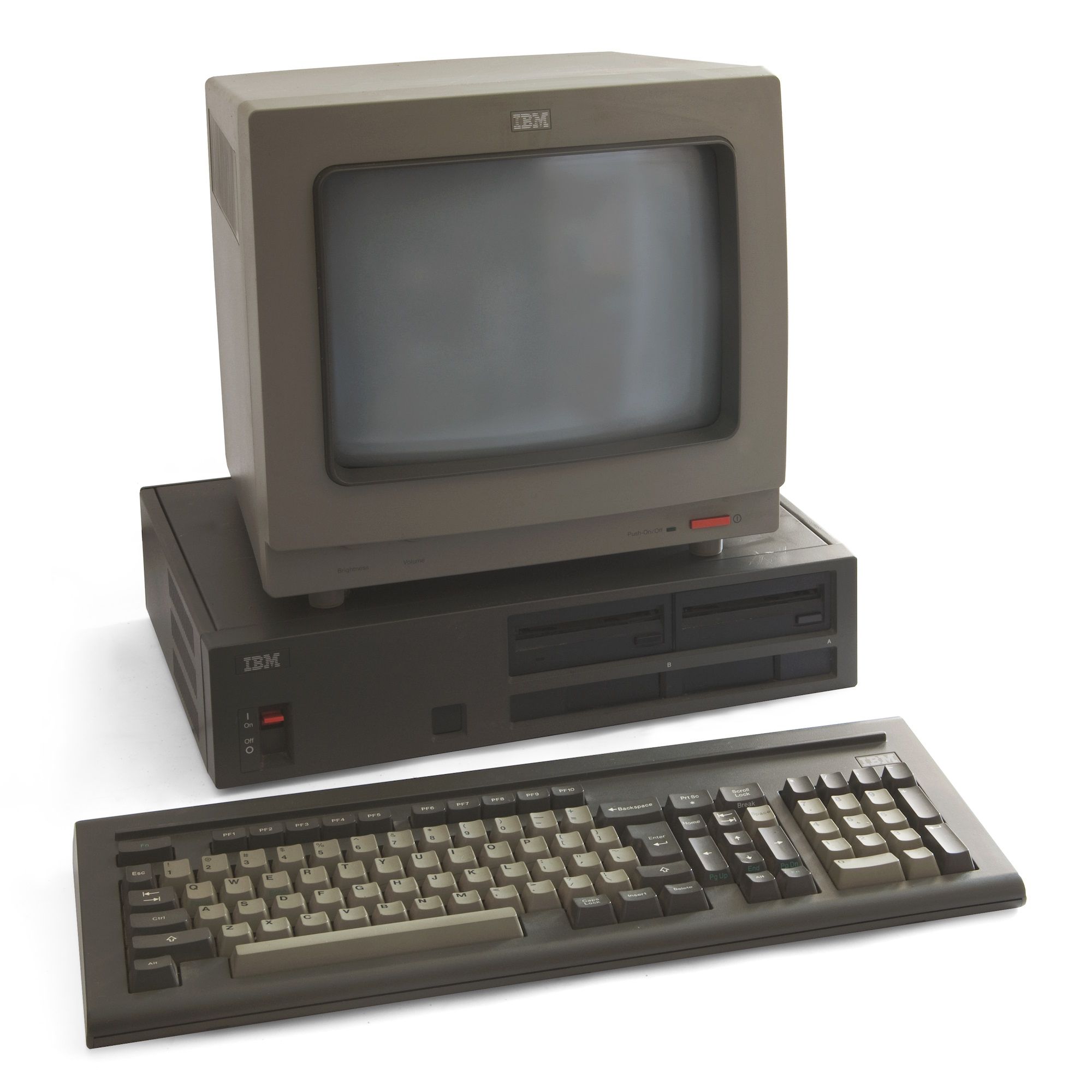 A three-piece desktop computer.  Up top is a black CRT, with a single red power button.  The pizzabox style case features two 3.25" drives, a recessed red power switch, and an unlabeled future expansion port.  The front of the case seems to slide off entirely, which would allow the top to hinge up and off, like their competitor Apple.  The keyboard, itself, is black in the familiar IBM layout, with one addition:  a raised ridge, where a student might rest a pencil.