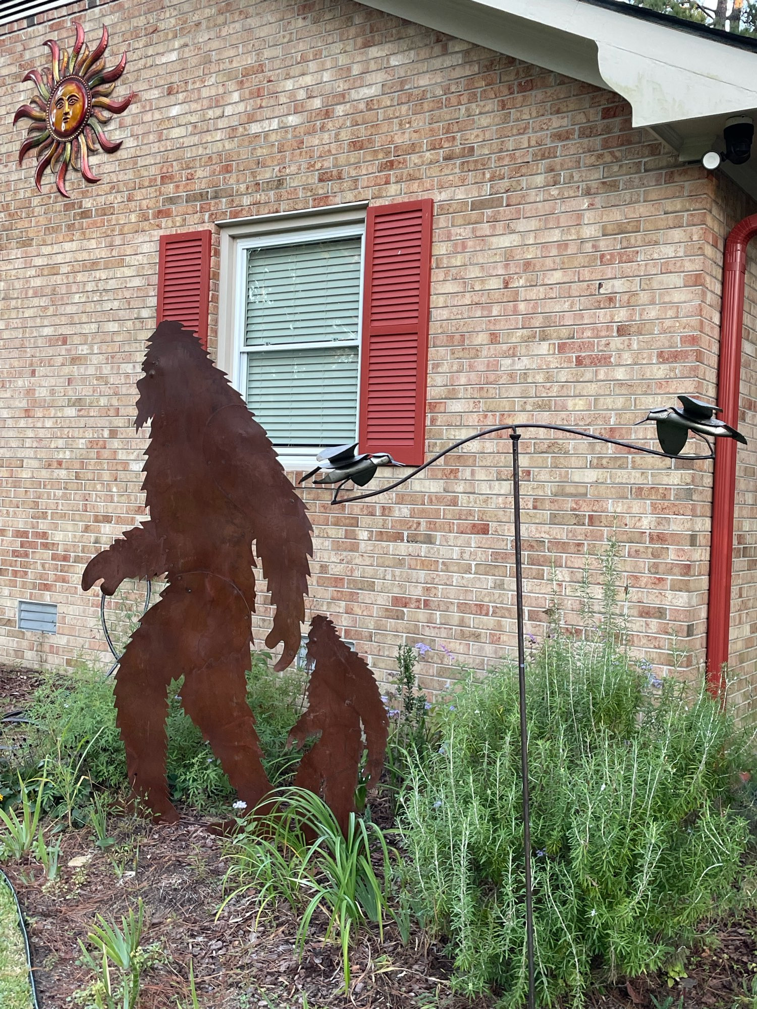 A Sasquatch family walks by front of our home. In foreground is a pivoting bird whirligig. Sun ornament graces the apex of the house wing. 