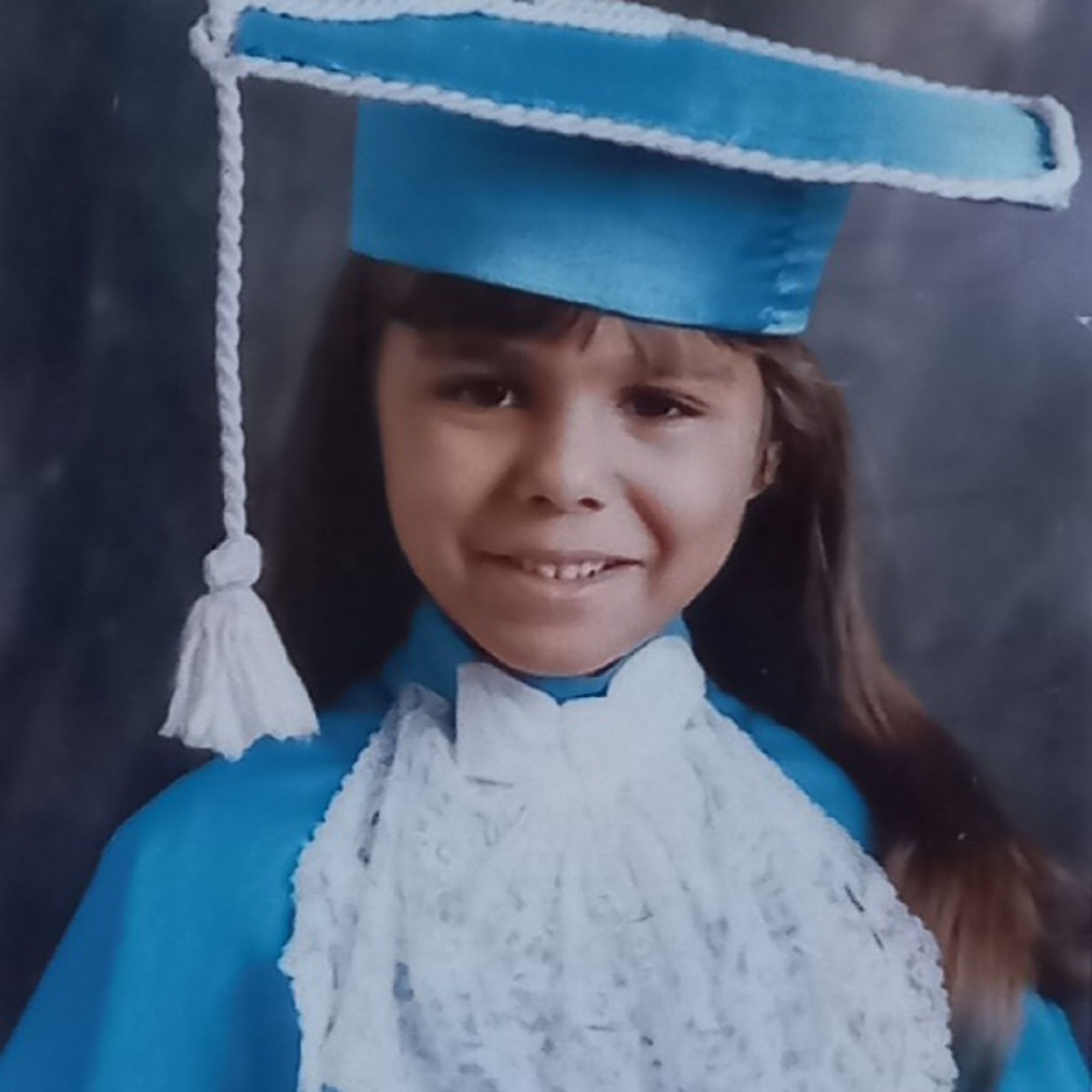 sorrindo com os dentinho de leite meio torto, cabelo solto e comprido com franjinha, beca e chapéu de formanda na cor azul clara com detalhes em branco