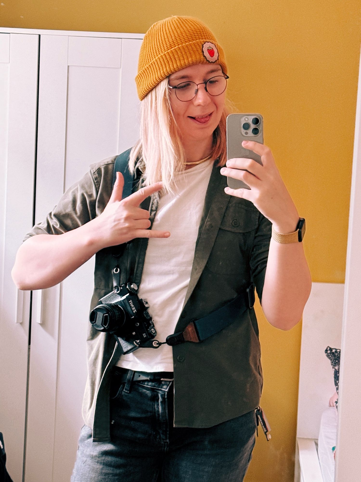 maisie making the horns sign. she’s wearing a white shirt with green over shirt and a yellow beanie. a zenit film camera is strapped to its body