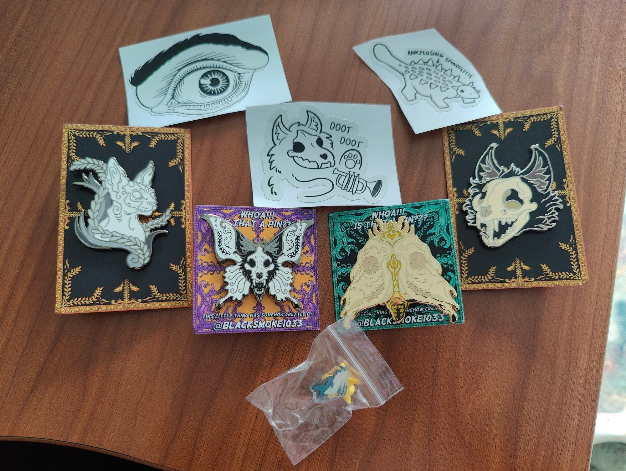 A selection of pins and stickers. The stickers are monochrome, with one highly detailed eye, one sketchy skulldog with a trumpet and the words 'doot doot' and one doodle of an Ankylosaurus labelled 'ankylosing spondylitis'. The pins are of a sphinx cat, a skullcat, and two different designs of lepidoptera incorporating animal skulls.