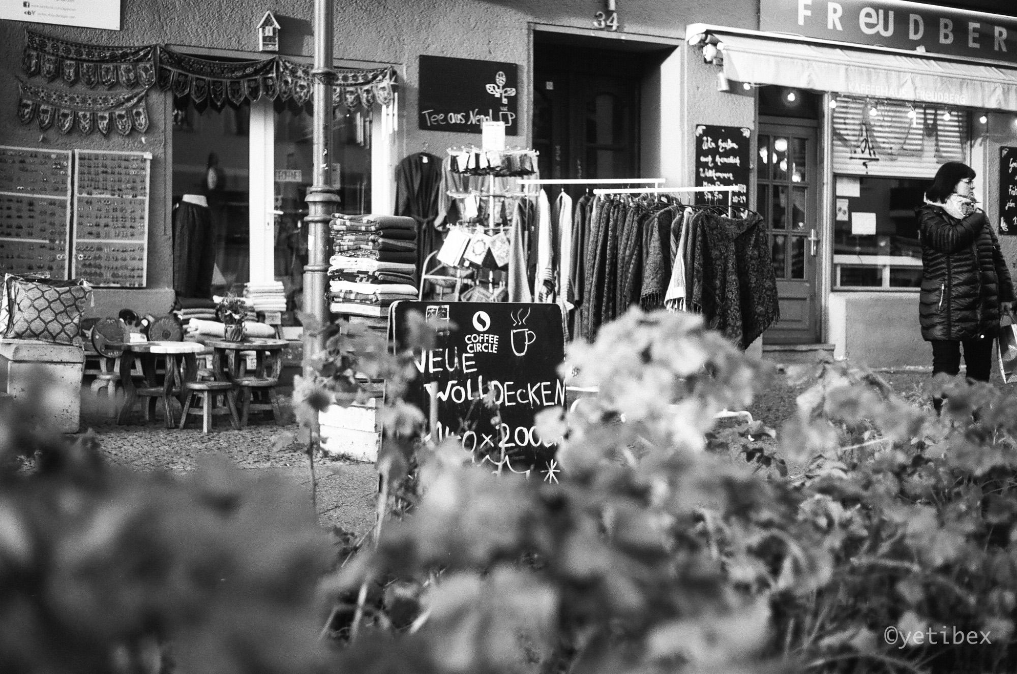 A street scene from Berlin: Looking over a hedge at some shoppes, their wares put out onto the sidewalk for display. A placard reads, "Neue Wolldecken," which translates to "new wool blankets."