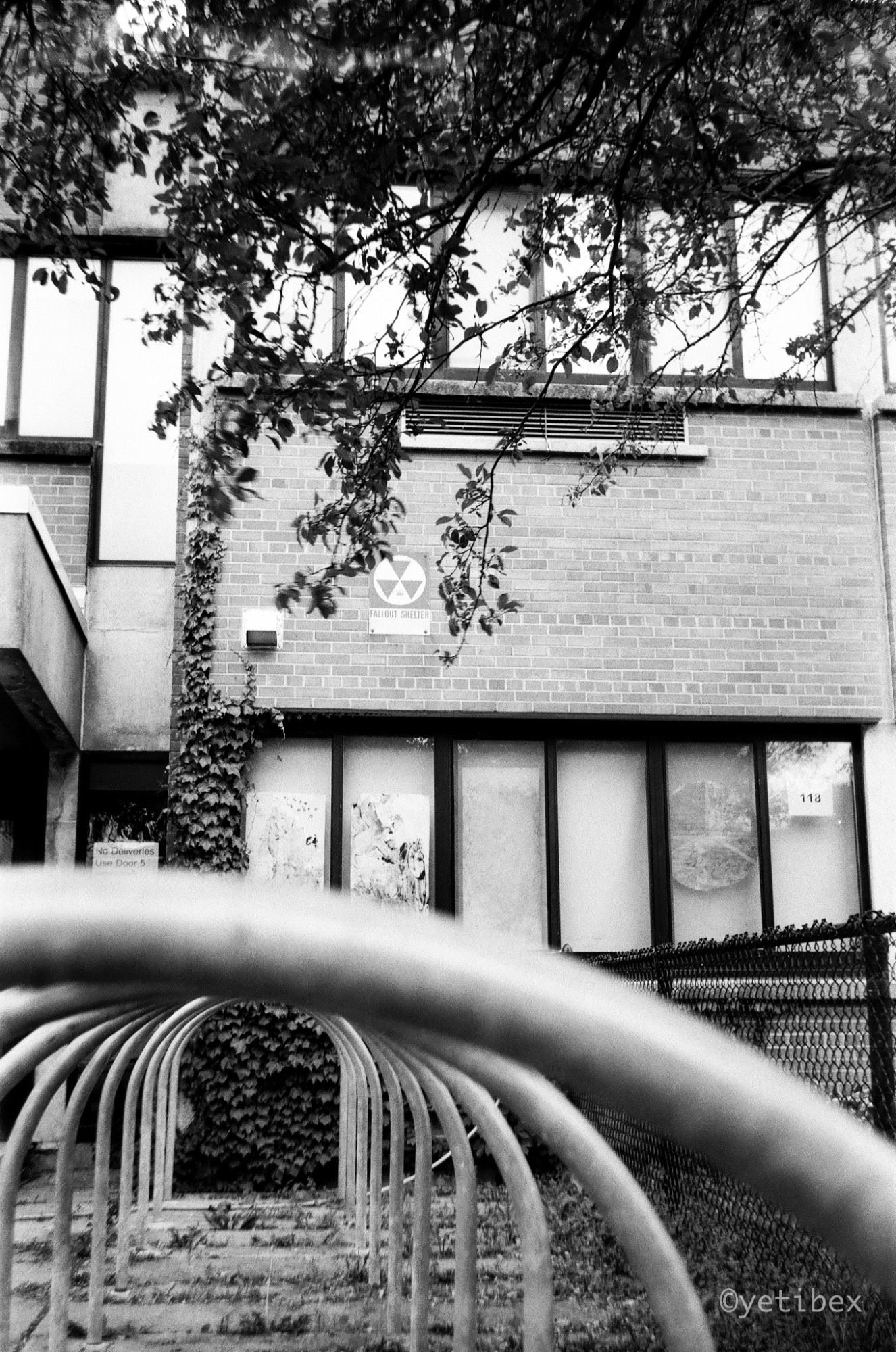 A black and white image of a school building. The lower part of the image is shot through a bicycle rack which seems to form a tunnel. The sign on the building says, "Fallout Shelter".