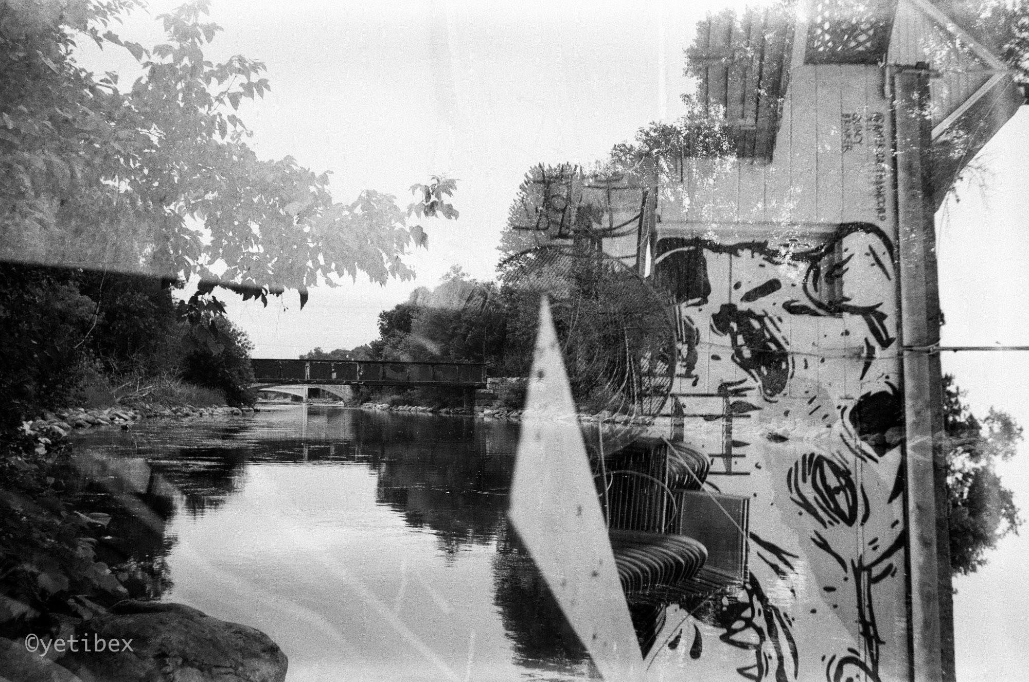 A black and white image shot as a multiple exposure, where the left side shows a view down a river towards a bridge, while the right side of the image juxtaposes - at a 90º angle - a graffiti painted building.