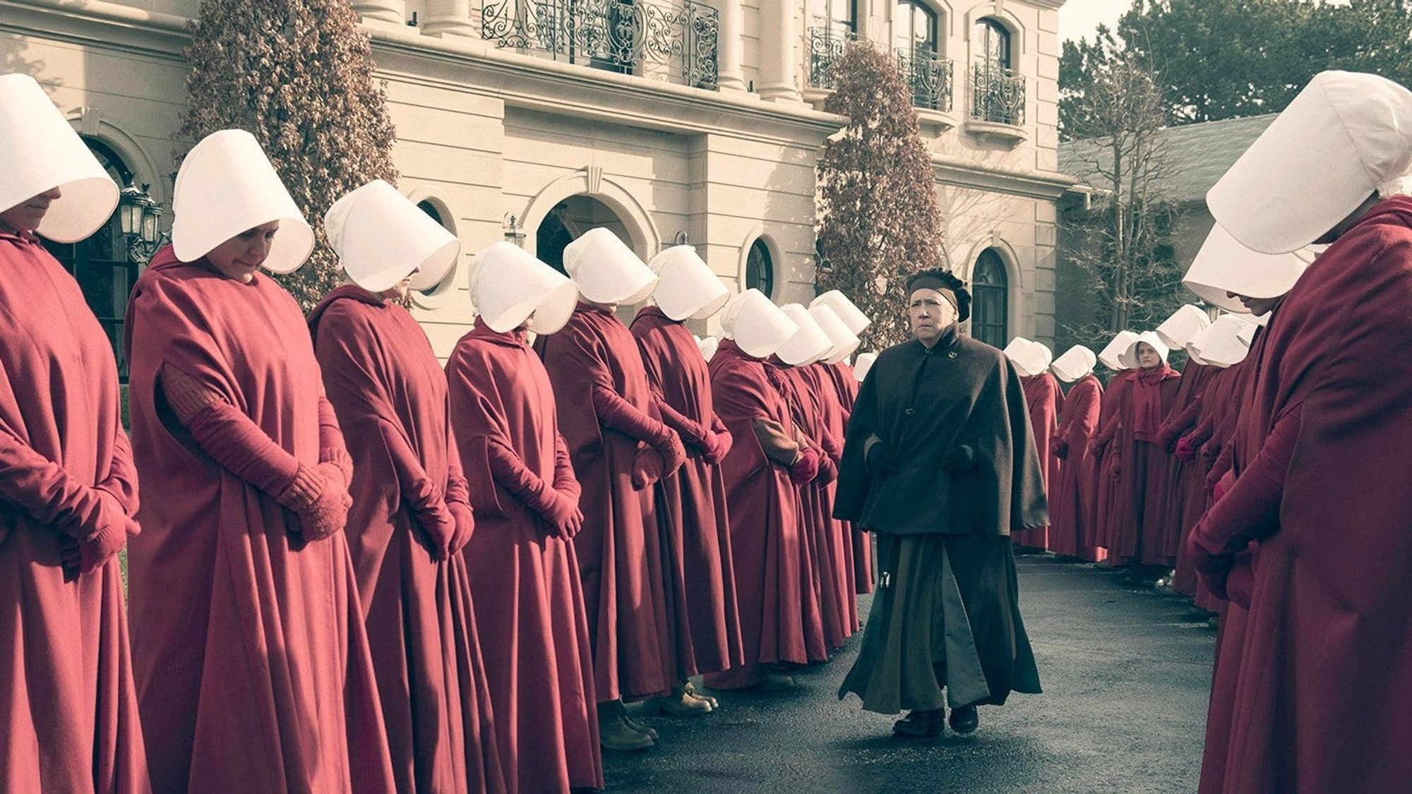 A group of women in red robes and white bonnets are standing in a line. At the front of the line, a woman in a dark brown coat and hat is walking. The women are Handmaidens, the female servants in the dystopian society depicted in the TV show "The Handmaid's Tale." The image captures the starkness and conformity of the society, as well as the power dynamics between the Handmaids and the ruling class.