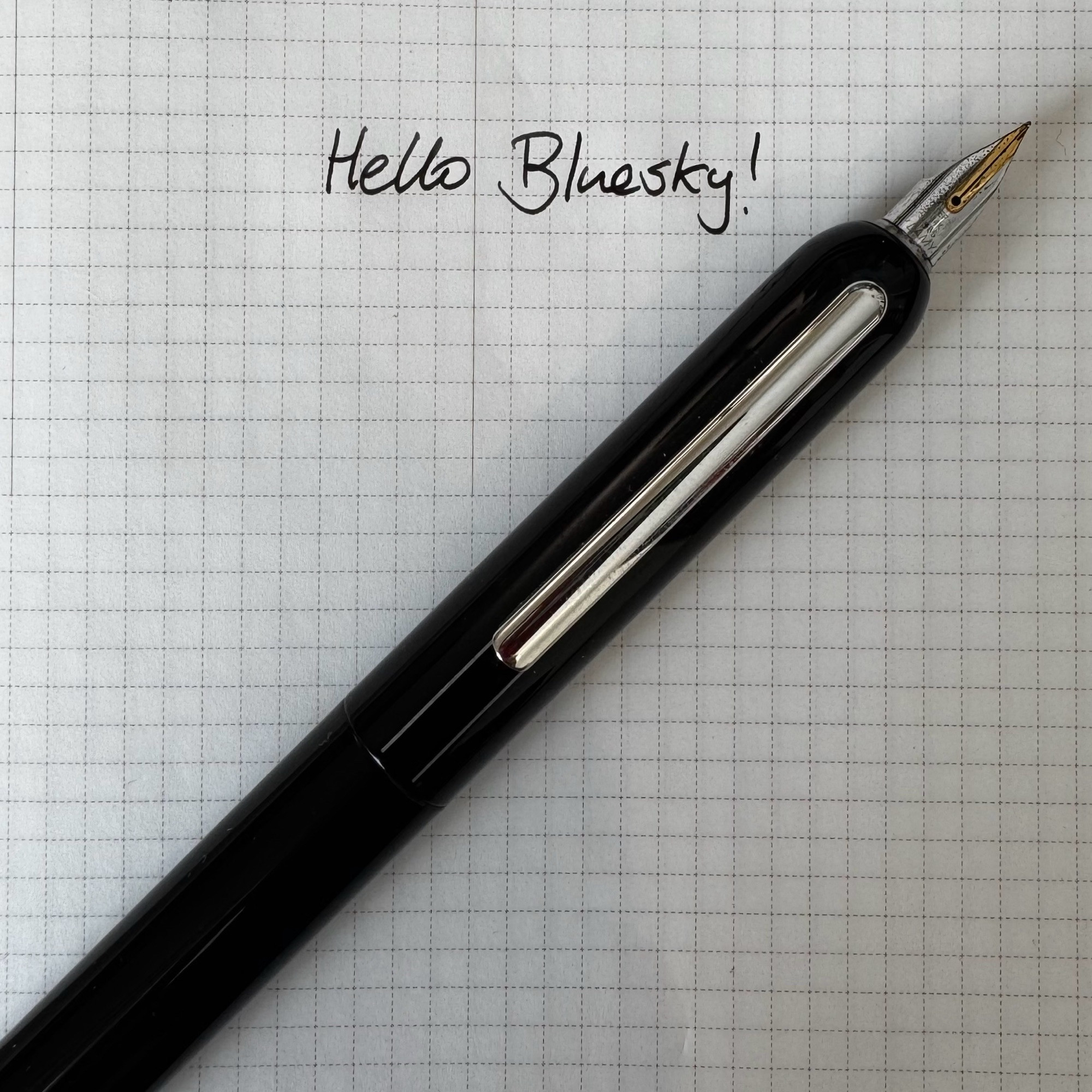 A piano black fountain pen sitting on grid paper, next to a handwritten message that says “Hello Bluesky!”.