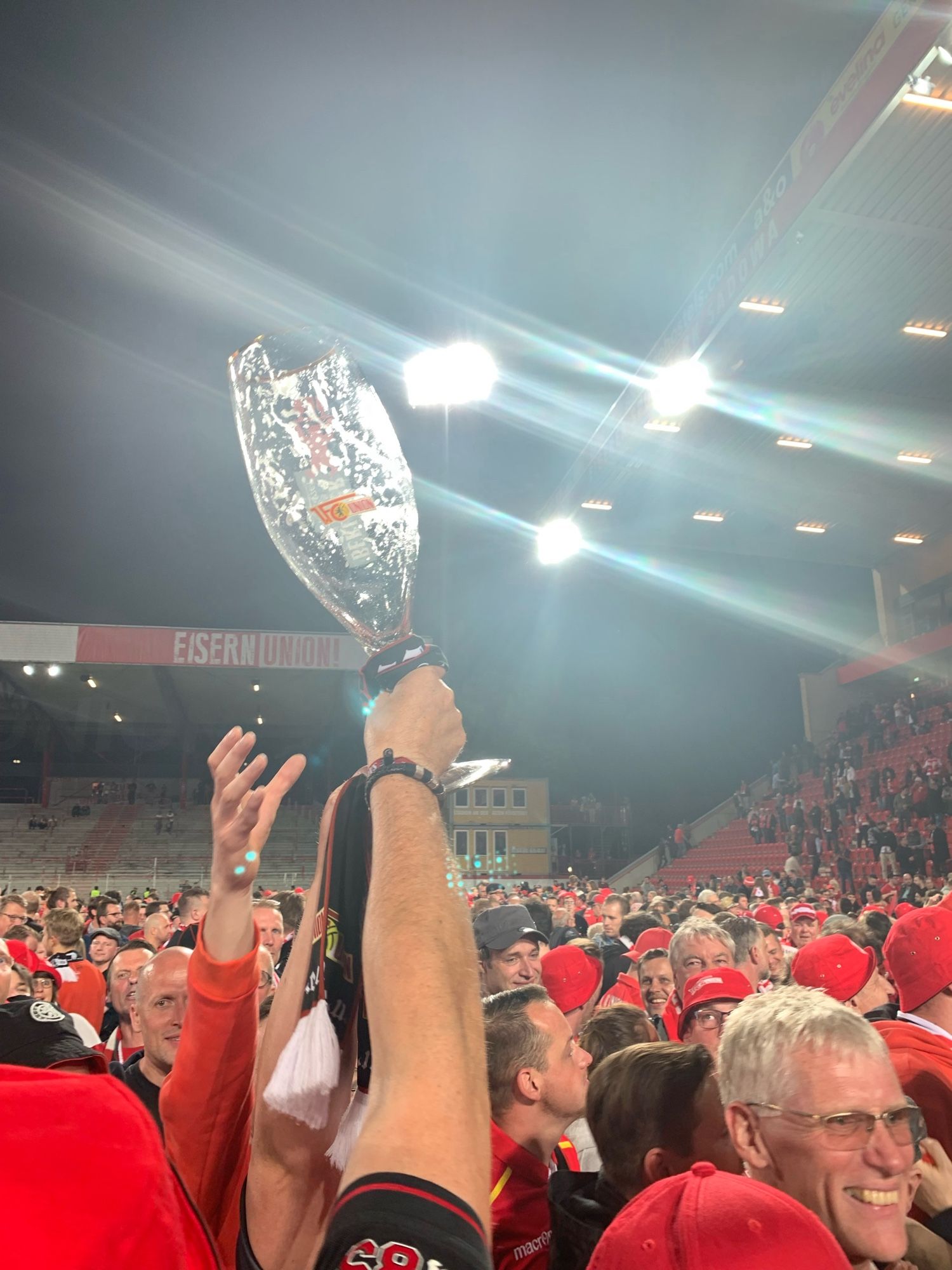 Großes Bierglas als Trophäe in den Himmel gestreckt in einem Pulk Menschen auf einem Fußballplatz.