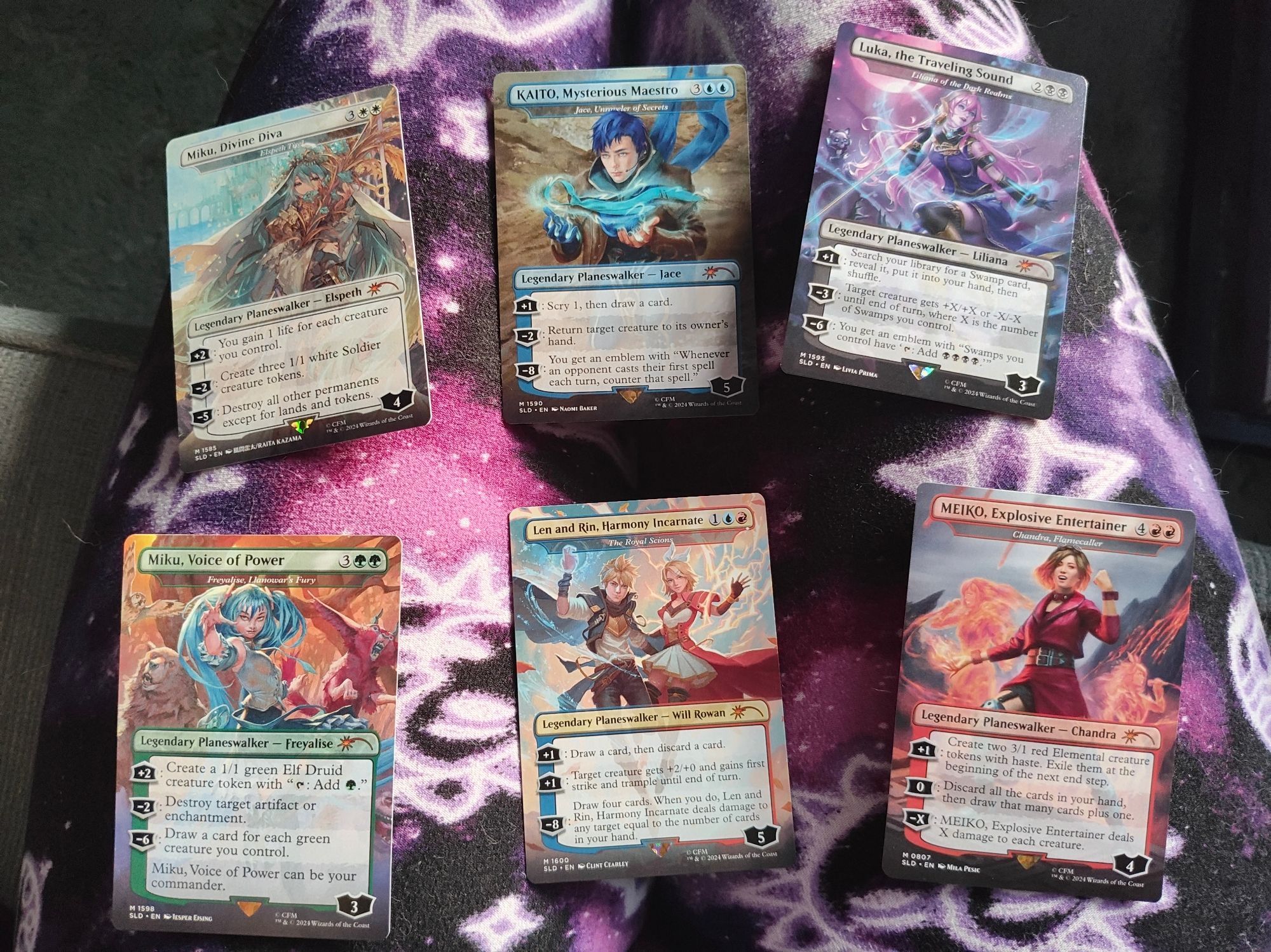 Six Magic the Gathering cards laid out upon a lap. From left to right, top to bottom, the cards are as follows:
Miku, Divine Diva (Elspeth Tirel)
KAITO, Mysterious Maestro (Jace, Unraveler of Secrets)
Luka, the Traveling Sound (Liliana of the Dark Realms)
Miku, Voice of Power (Freyalise, Llanowar's Fury)
Len and Rin, Harmony Incarnate (The Royal Scions)
MEIKO, Explosive Entertainer (Chandra, Flamecaller)