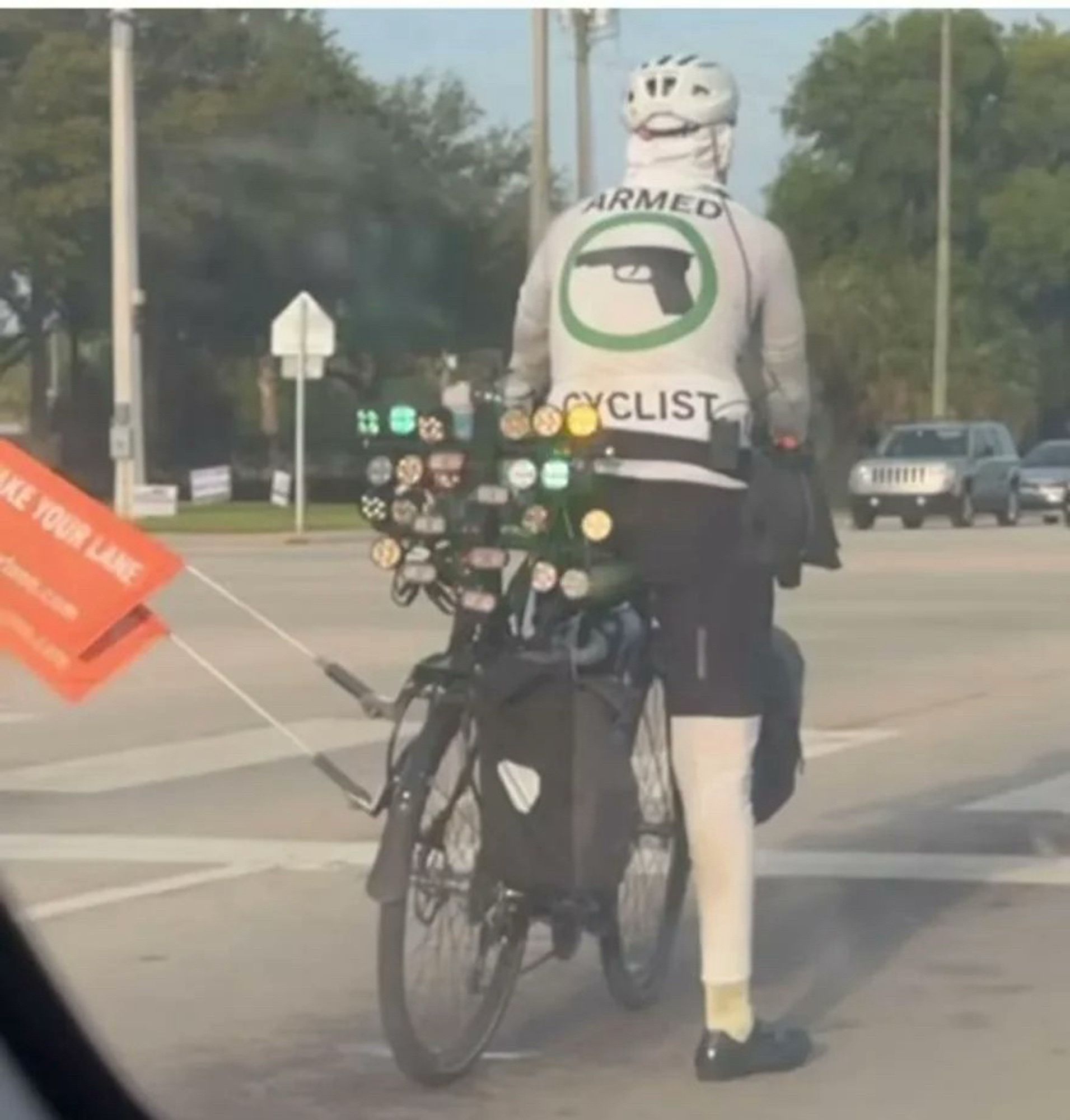 Militanter Kampfradler an einer Straßenkreuzung. Auf seinem T-Shirt steht „Armed Cyclist“. An seinem Fahrrrad sind zahlreiche Rückleuchten angebracht, sowie mehrere seitlich angebrachte Fahnen, welche die Einhaltung des Sicherheitsabstands beim Überholen anmahnen