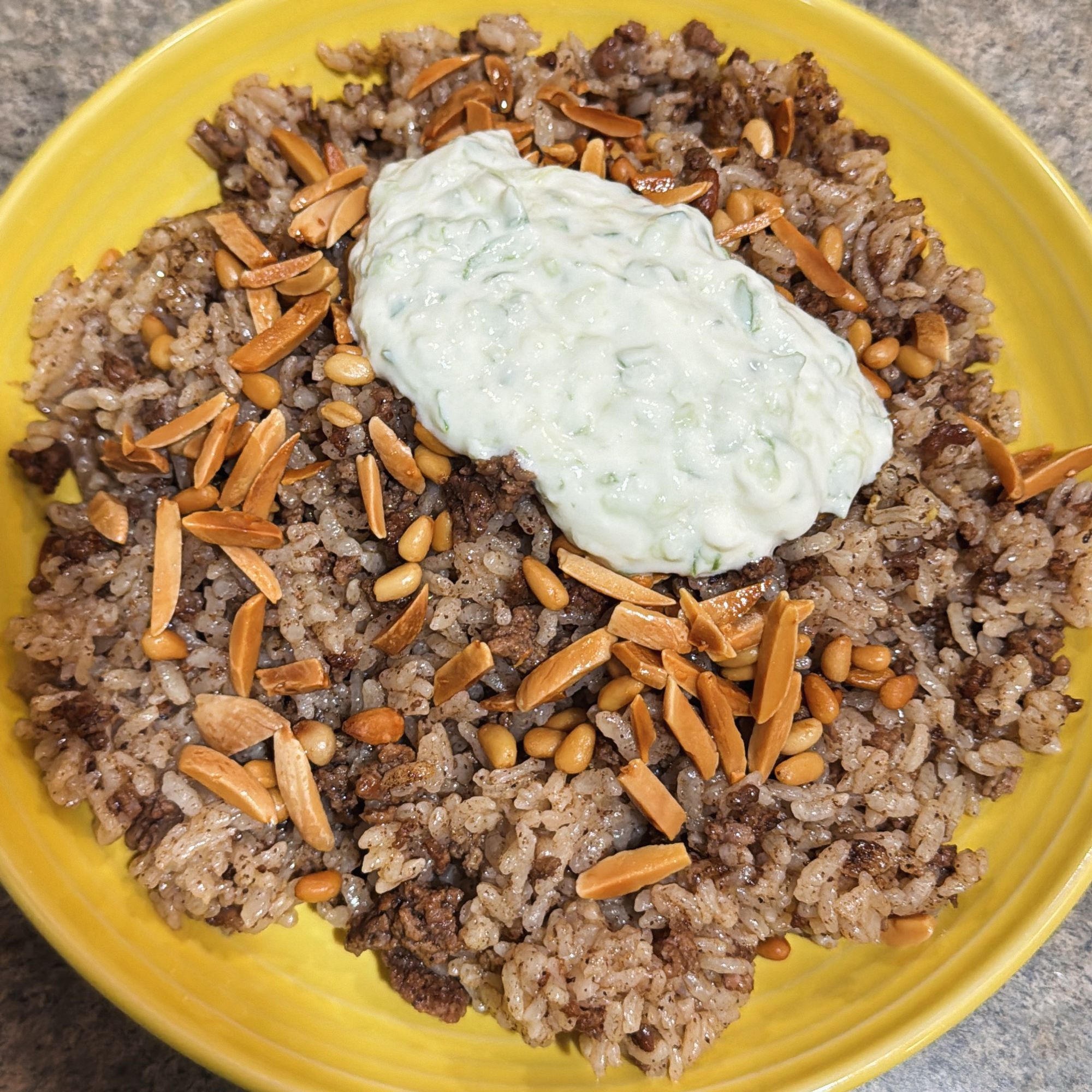 A yellow fiesta bowl plate filled with hashweh, a spiced beef-rice dish topped with toasted pine nuts & slivered almonds, and a dollop of tzatziki.