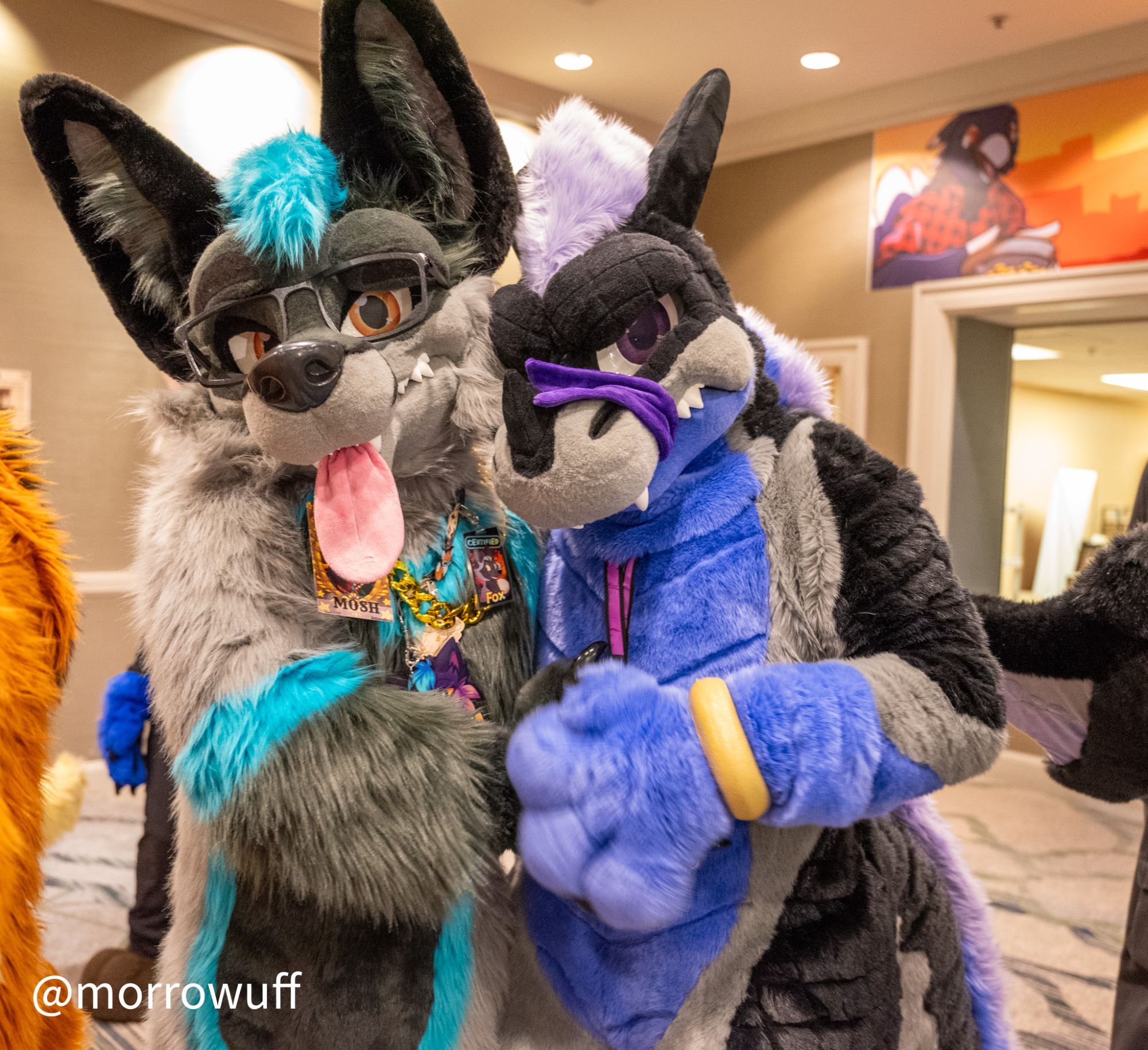 DenFur 2023. Photo of two large and cartoony-styled More Fur Less fursuiters: Mosh, a gray fox with blue markings; and Darris, a purple, gray, and black dragon.