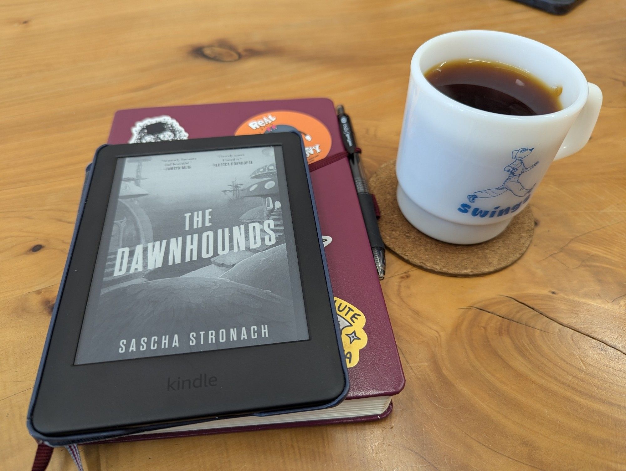 A Kindle displaying the cover to 'The Dawnhounds' by Sascha Stronach, a notebook covered in stickers, and a mug of coffee sitting on a wooden tabletop.