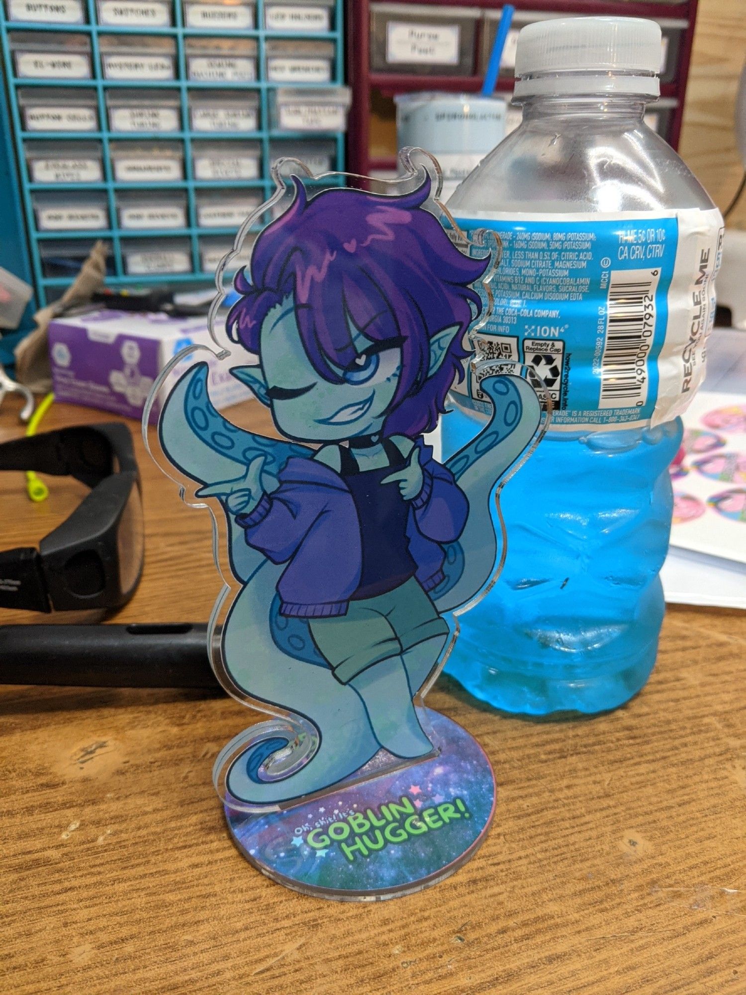 A home made Acrylic Standee of Goblinhugger, they are winking and shooting finger guns, they are green, with purple hair, and have 3 tenta coming out from behind them.