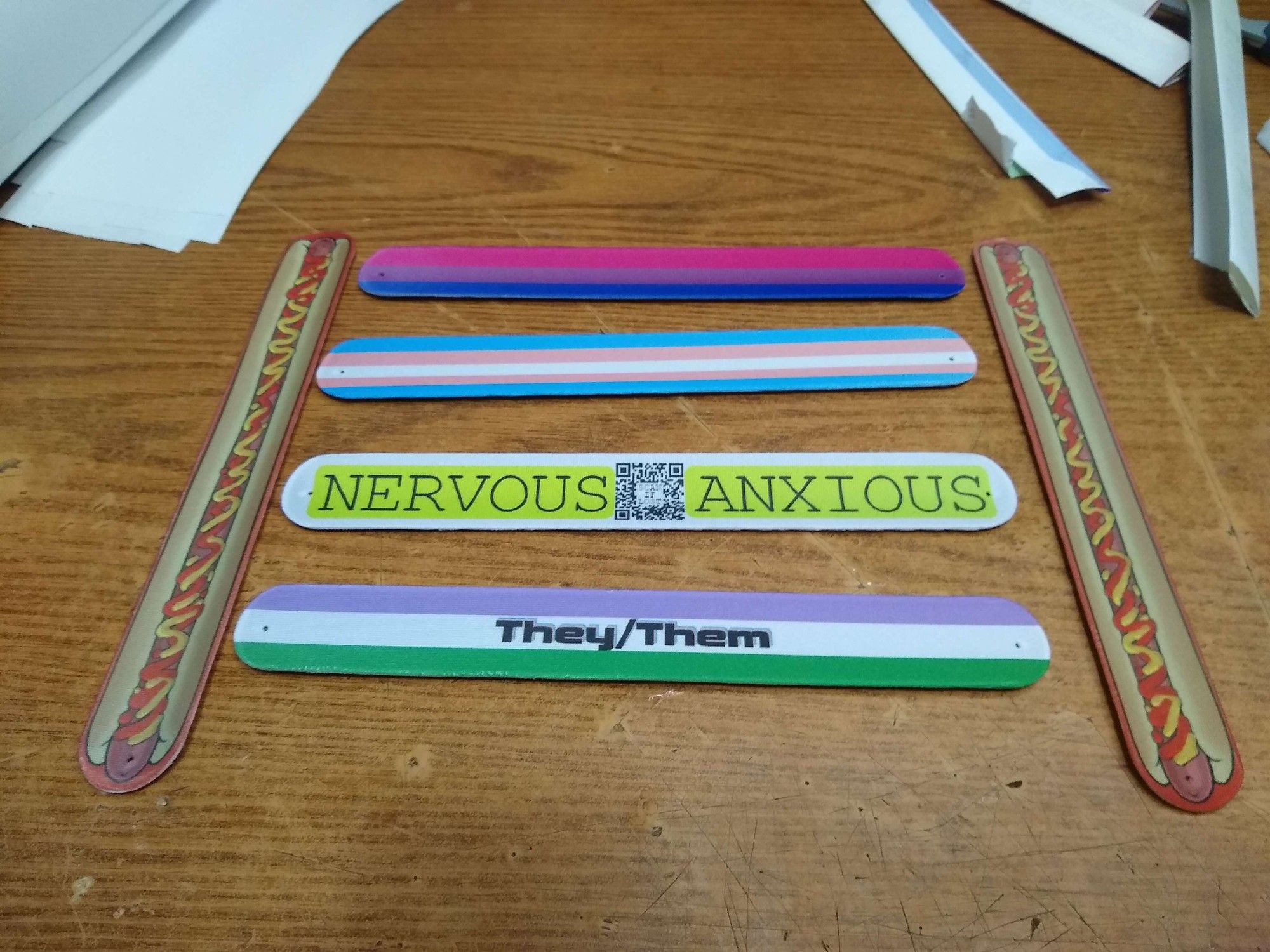 An assortment of Slap bracelets, 2 with Hotdog Prints, A Non Binary with They/Them Written on it, another with NERVOUS / ANXIOUS with a QR Code, one with the TRansFlag Colors and another with Bi Flag colors