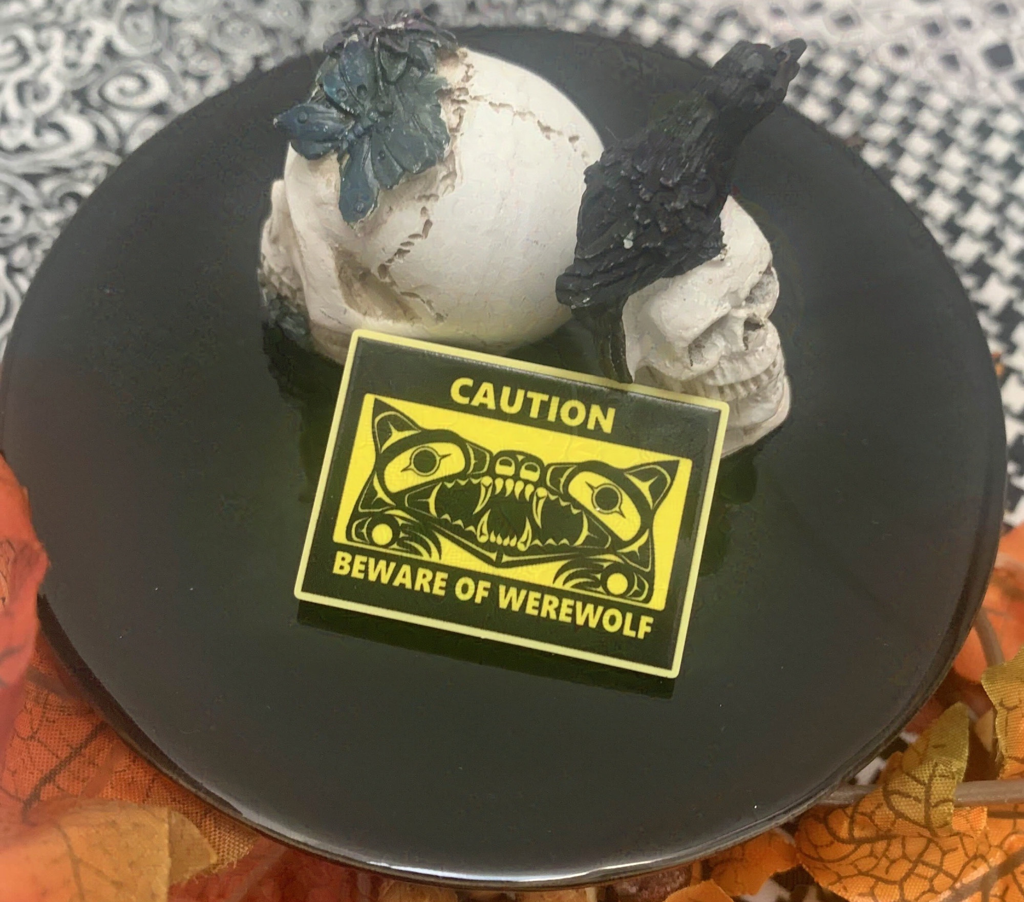 A yellow acrylic pin in the style of a warning sign with a formline werewolf on it in black.