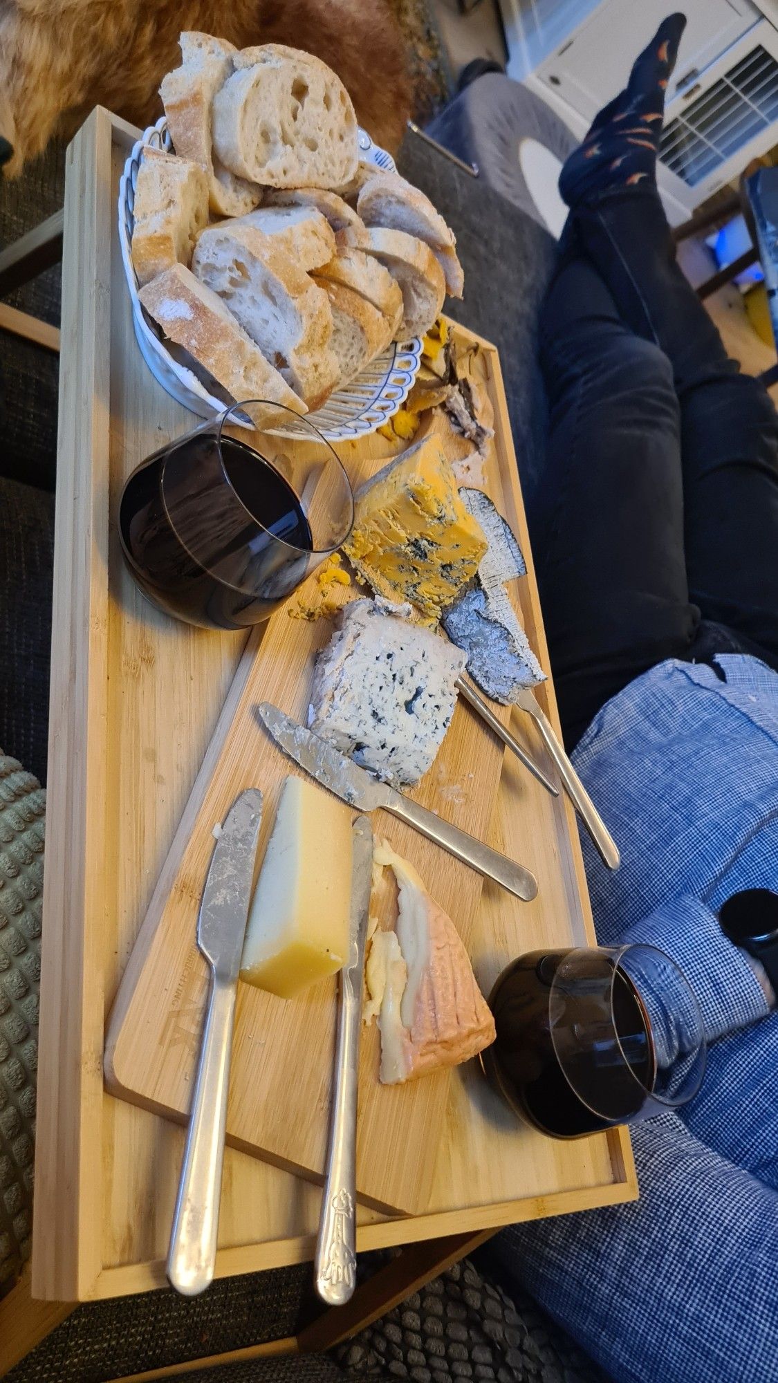 A delicious looking cheese board with two glasses of red wine and a basket of bread