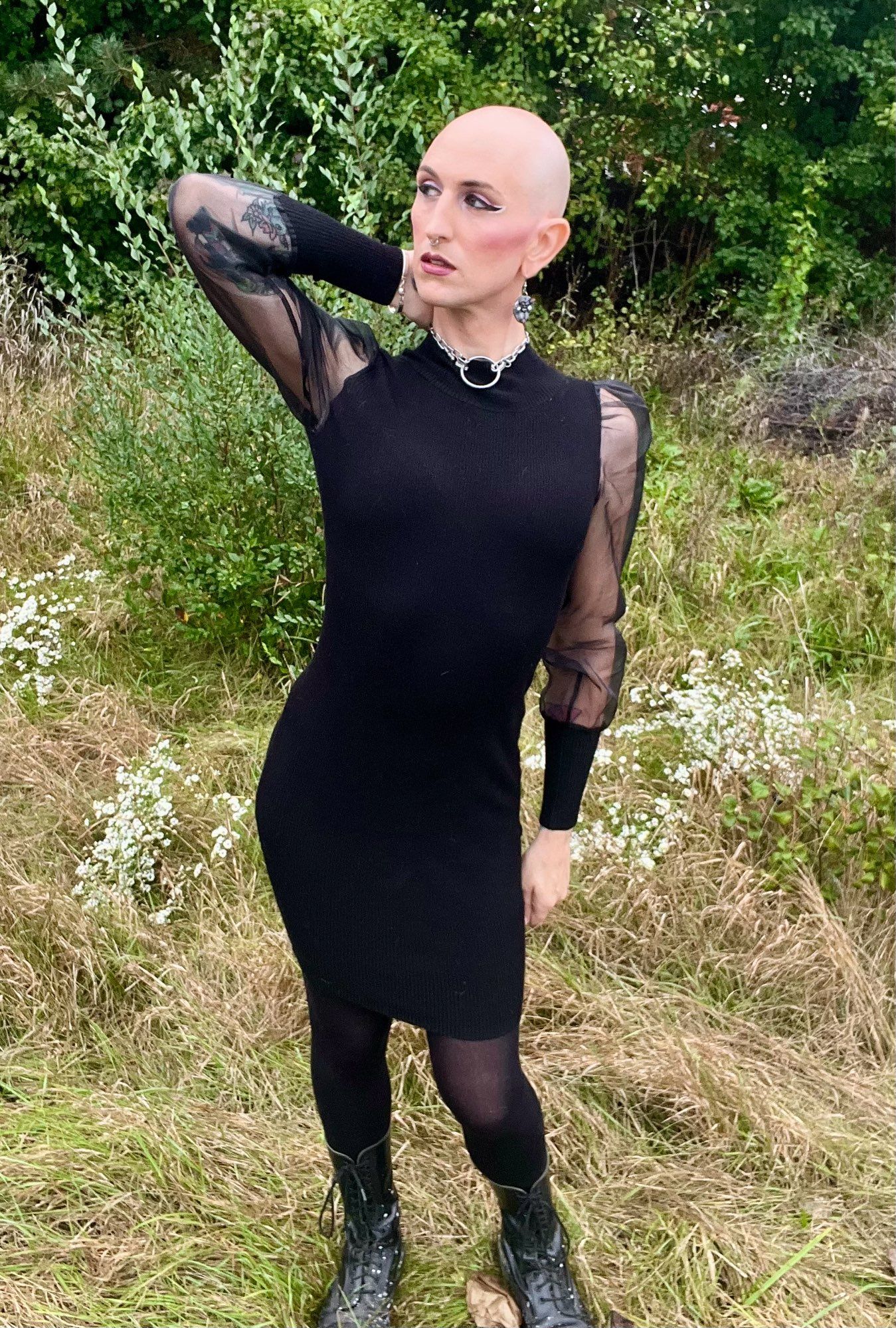 A bald femme in a black dress with semitransparent sleeves and black boots looking off frame and holding a dramatic pose