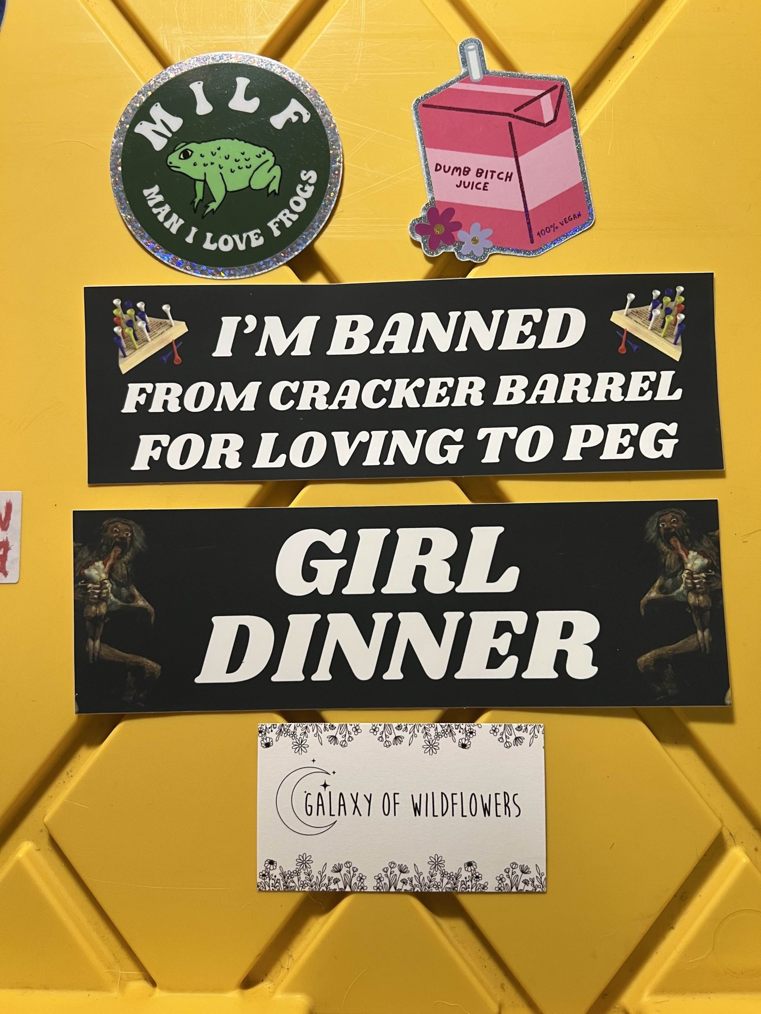 Four stickers sitting on top of a black and yellow tote:
1. A circular sticker depicting a green frog with the text "MILF - Man I Love Frogs"
2. A pink juice carton with pink and white flowers near the bottom. The juice box label reads "Dumb Bitch Juice"
3. A bumper sticker that reads "I'm banned from Cracker Barrel for loving to peg" with pictures of the peg game that's at all the tables
4. A bumper sticker depicting Saturn Devouring His Son that reads "GIRL DINNER"

At the bottom of the image is a business card for Galaxy of Wildflowers