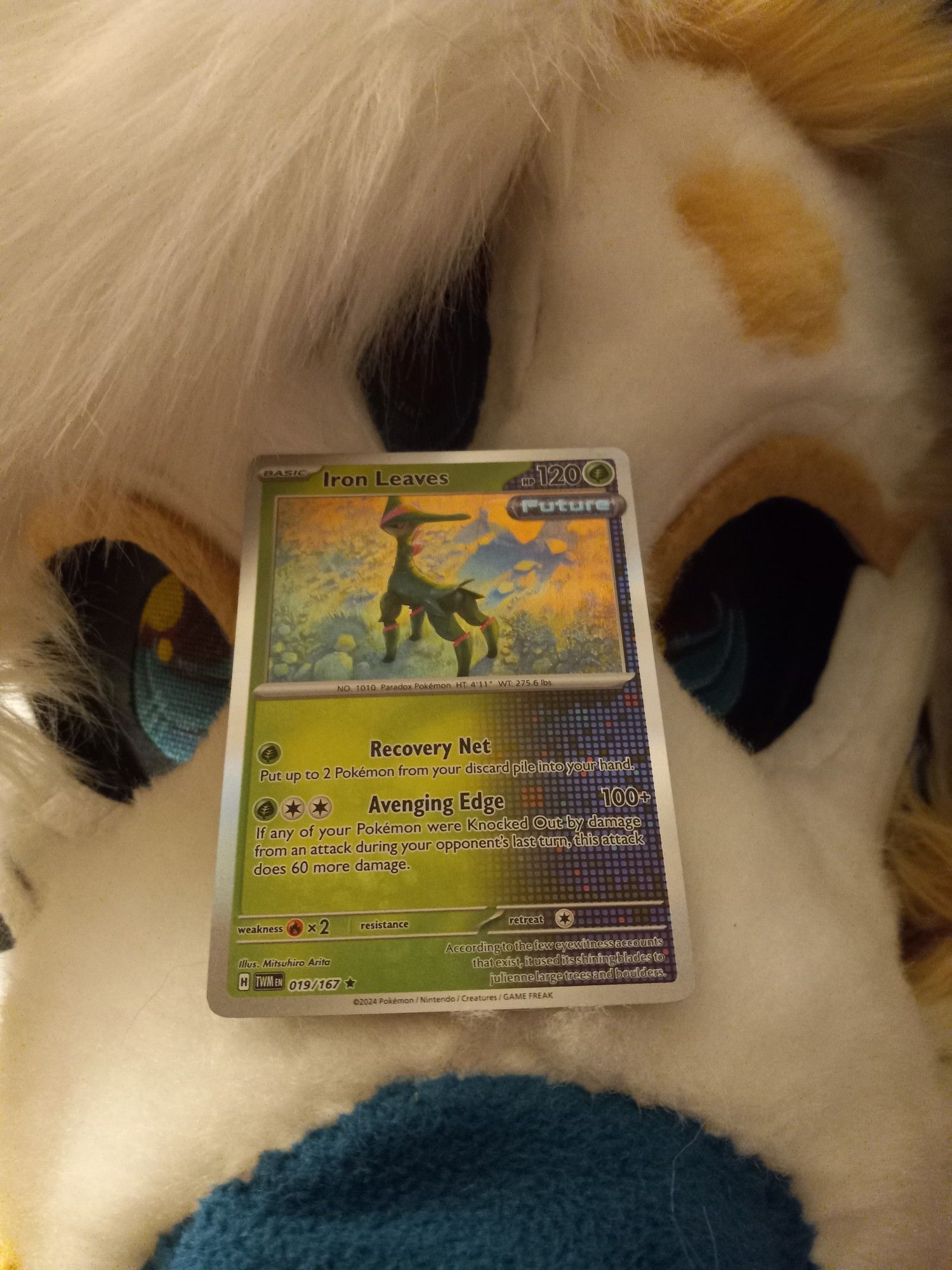 A picture of an Iron Leaves Pokemon card sitting on top of my fursuit's face.