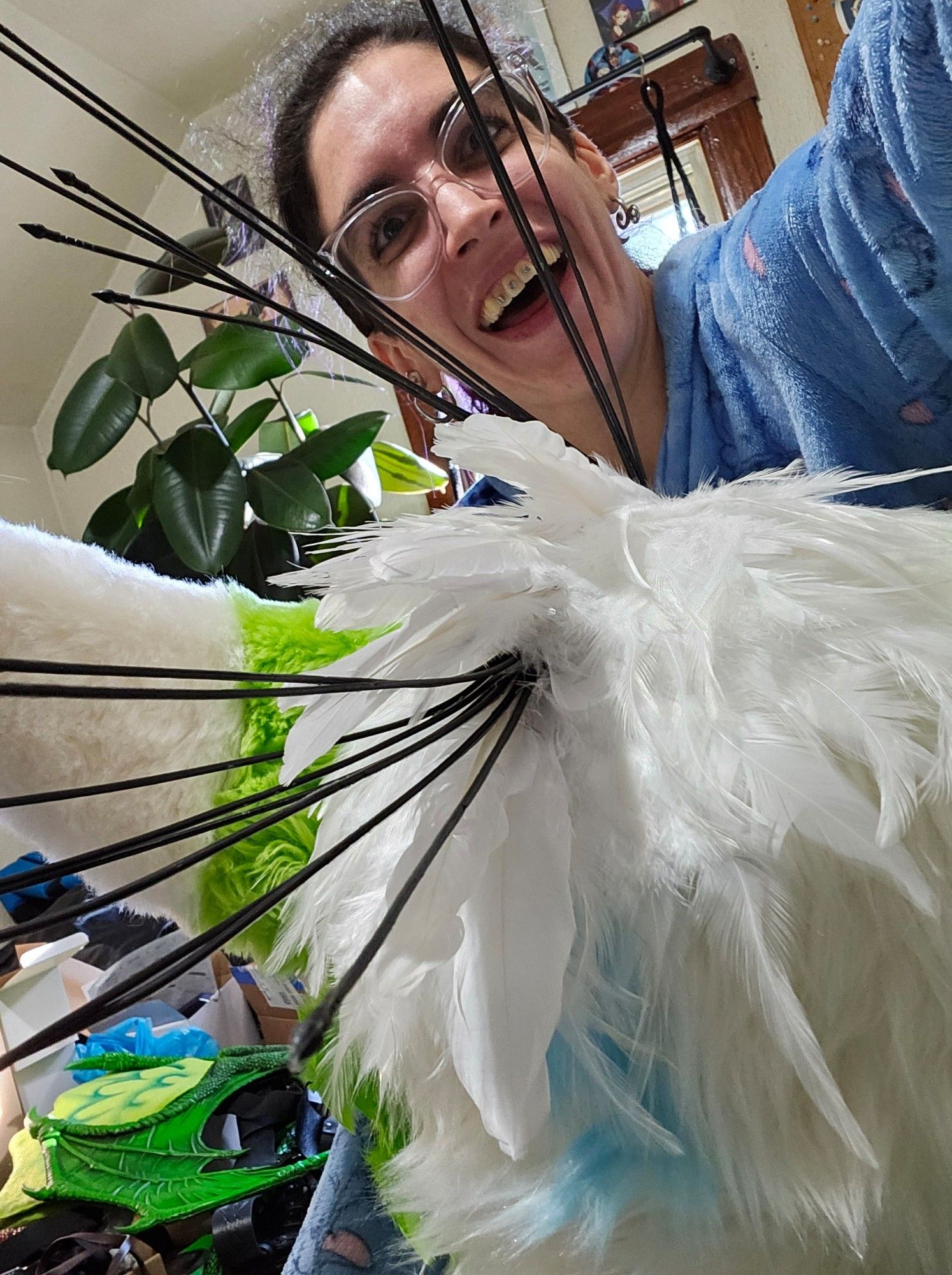 Me posing with my WIP feathering job on my gryphon fursuit head. A rubber plant and dragon wings are in the background lol