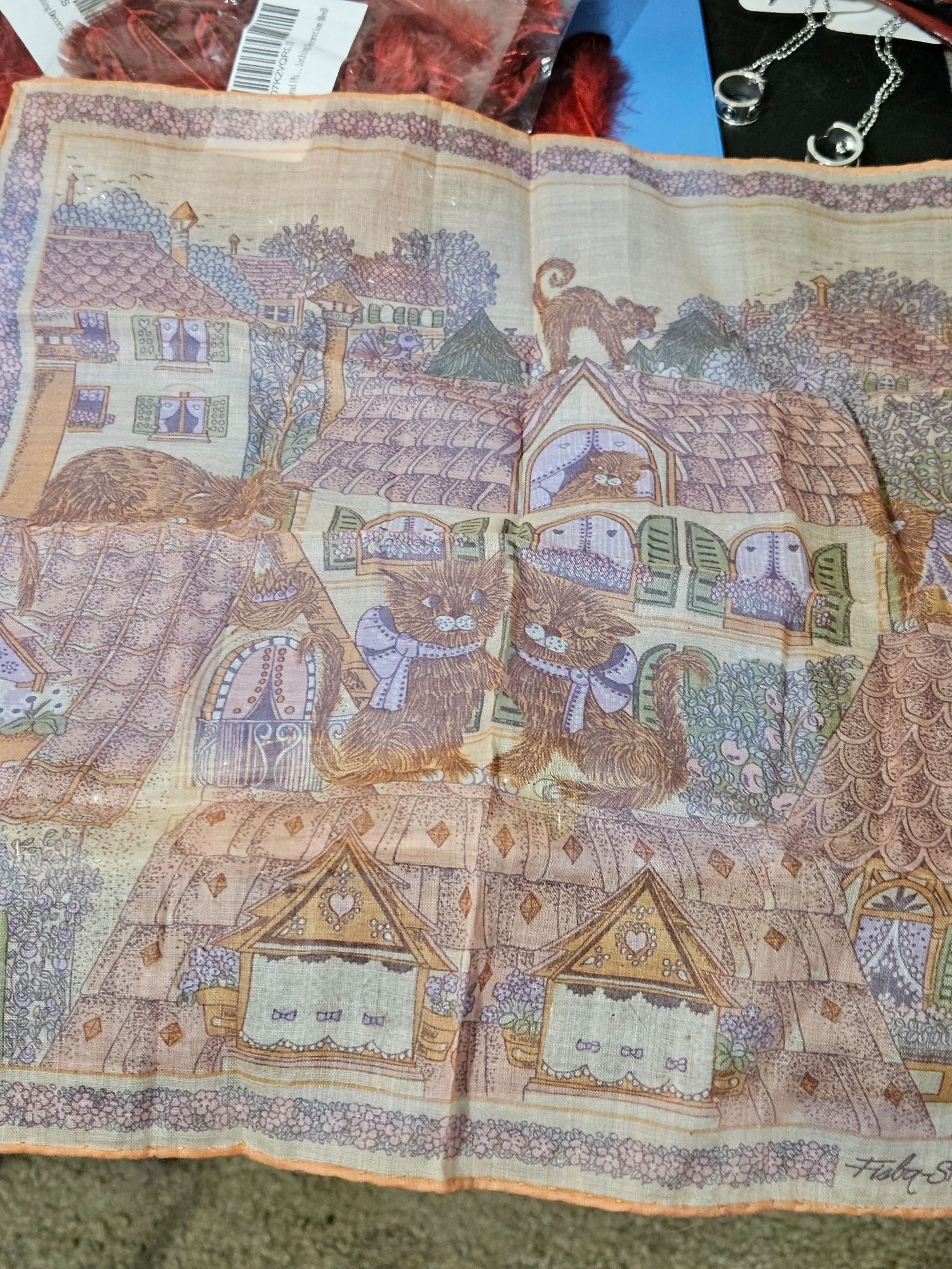 A vintage fisba stoffels handkerchief. Prolly silk, it's definitely fade, the over all colors are orange and yellow. It depicts cottage roofs with a pair of cats in the middle; sitting with the one on the left giving the other a paw. There are a couple of cats in the background cottage.