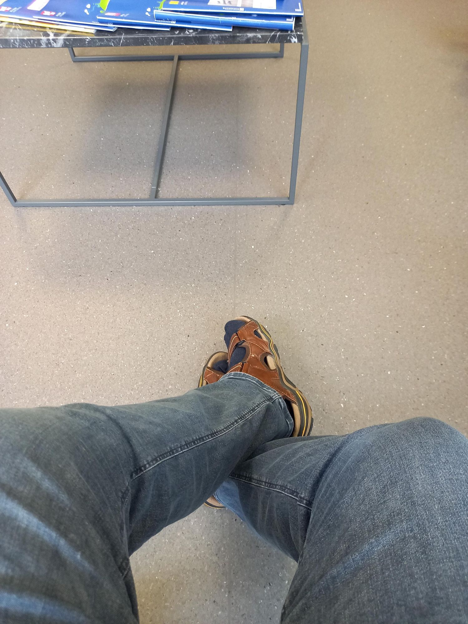 Im Vordergrund 2 Beine mit blauen Jeans und braunen Sandalen auf einem PVC-Boden, im Hintergrund das Metallgestell eines Tisches auf dem im Anschnitt noch blauen Schonumschläge eines Lesezirkel liegen