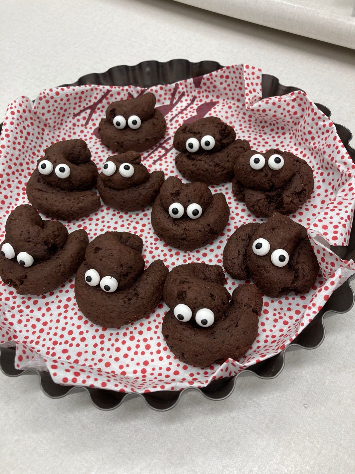 Auf einem mit einer weißen, rotgepunkteten Serviette ausgelegten Tablett liegen mehrere  braune Kekse mit Zuckeraugen. Die Kekse sind aus braunem, aufgerollten Teig und sehen auch wie Kackwürstchen