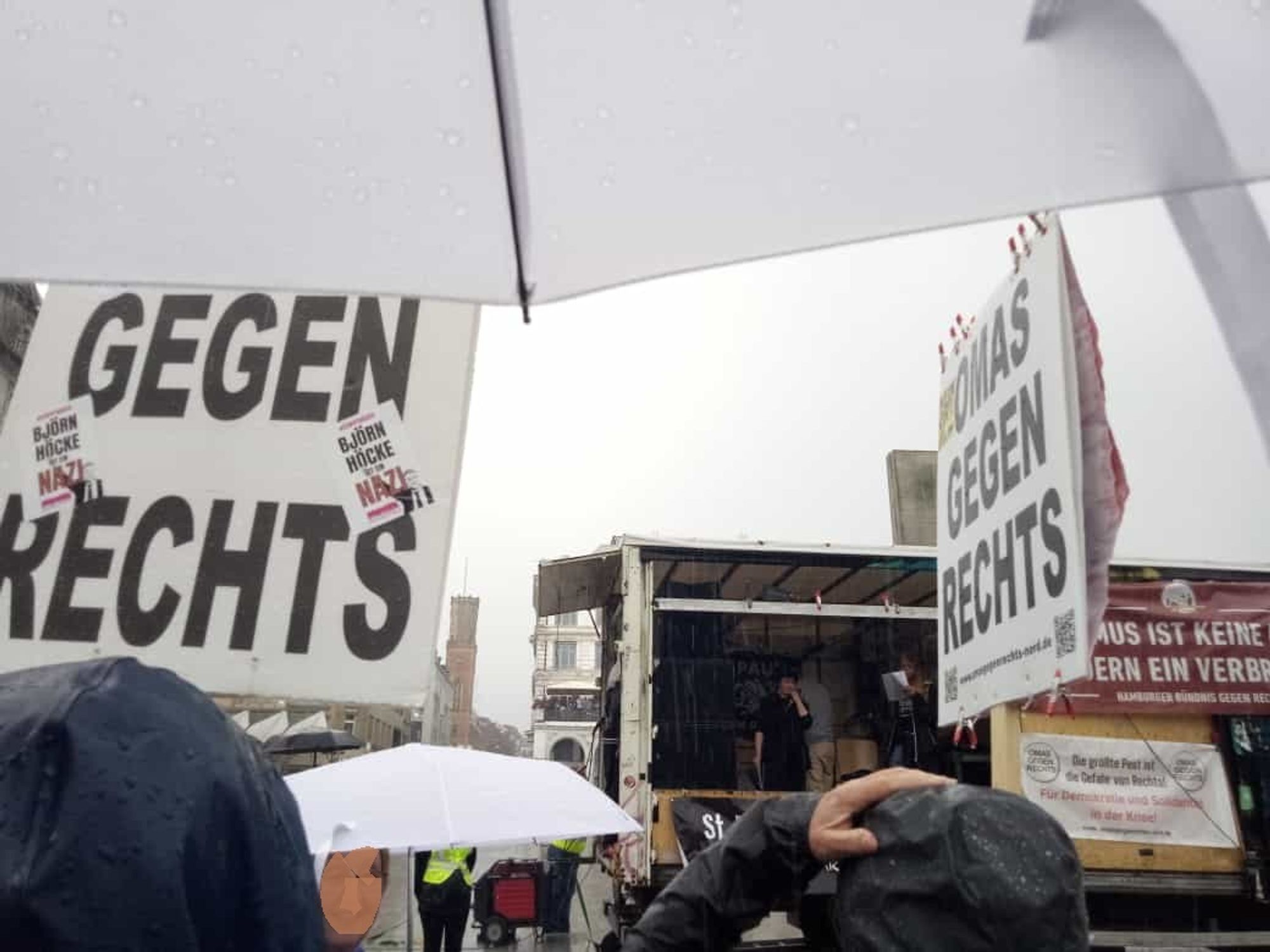 Blick auf den Lautsprecherwagen über ein paar Köpfe mit Kaputzen und einen Schirm. Schilder der OMAS GEGNE RECHTS sind zu sehen. Am Lauti hängt ein Banner des Hamburger Bündnis gegen Rechts "Faschismus ist keine Meinung sondern ein Verbrechen". Darunter ein Banner der OMAS "Die größte Peste ist die Gefahr von Rechts". Der Himmel ist weiß.