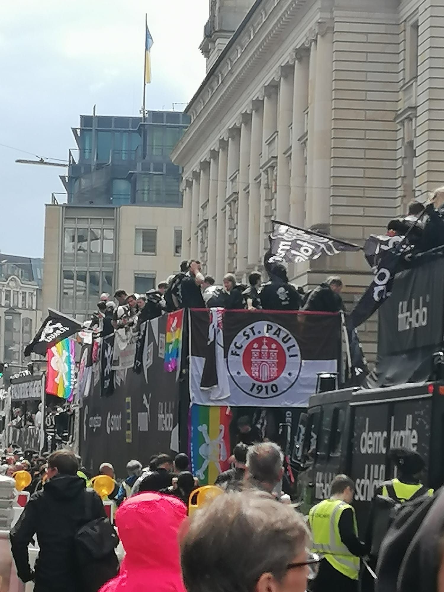 Zu sehen ist ein hoher Umzugswagen, auf dem oben die Mannschaft des FC St. Pauli steht. An der Rückseite hängt die Fahne des Vereins, an der Seite weitere Fahnen, dabei auch zwei in Regenbogenfarben. Dahinter folgt ein weiterer Umzugswagen und nebenher laufen viele Menschen. Die Wagen fahren an einer hohen Häuserzeile vorbei.